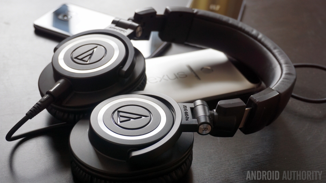 A pair of Audio Technica ATH-M50X wirred headphones resting flat on a table on top of three face-down smartphones.