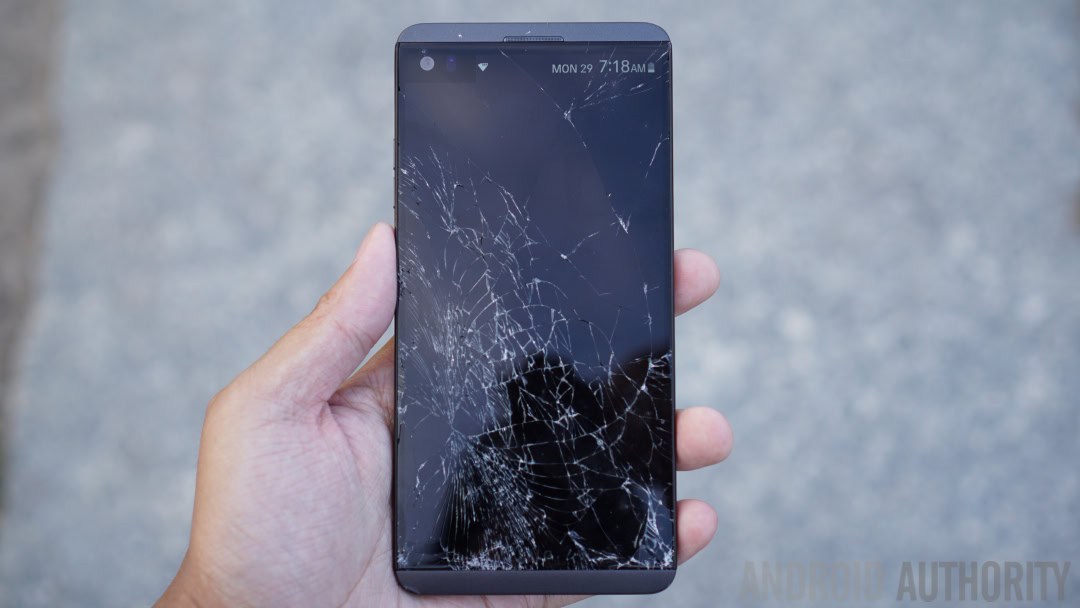 An LG V20 smartphone with a shattered screen held in a hand.
