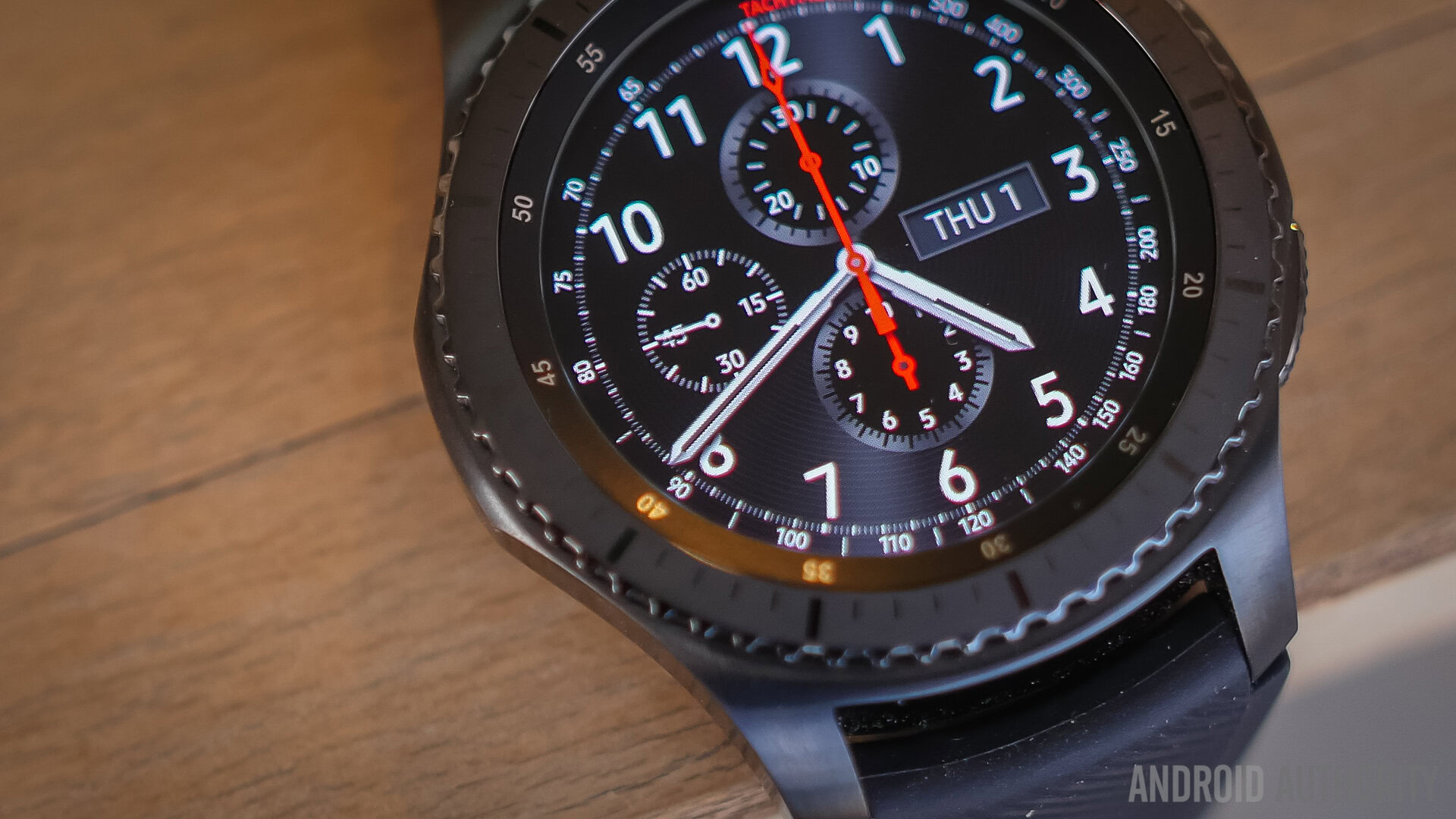 A Samsung Gear S3 rests on the edge of a wooden table. 