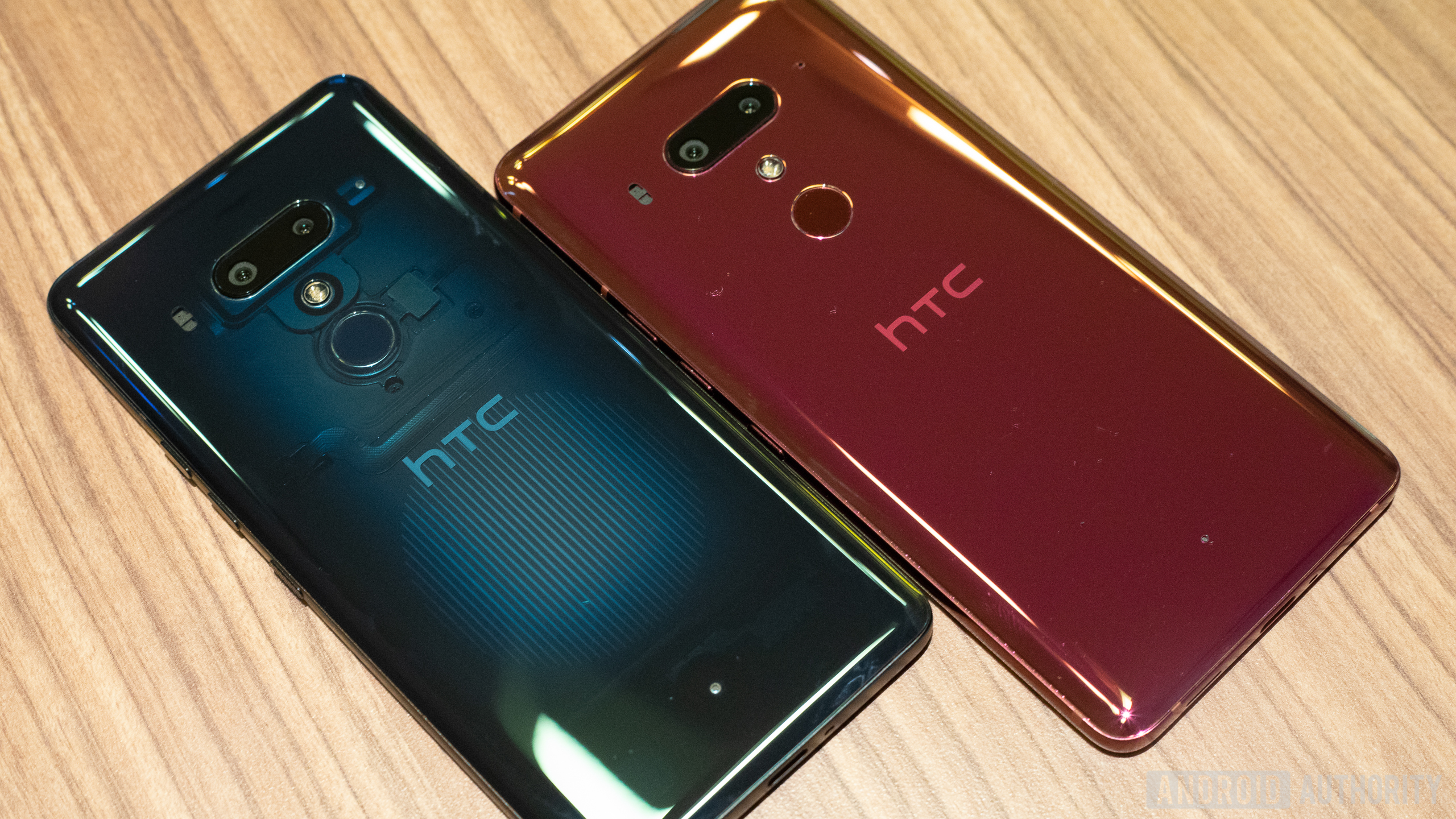 A blue HTC U12 Plus smartphone and a red HTC U12 Plus smartphone on a wooden surface.