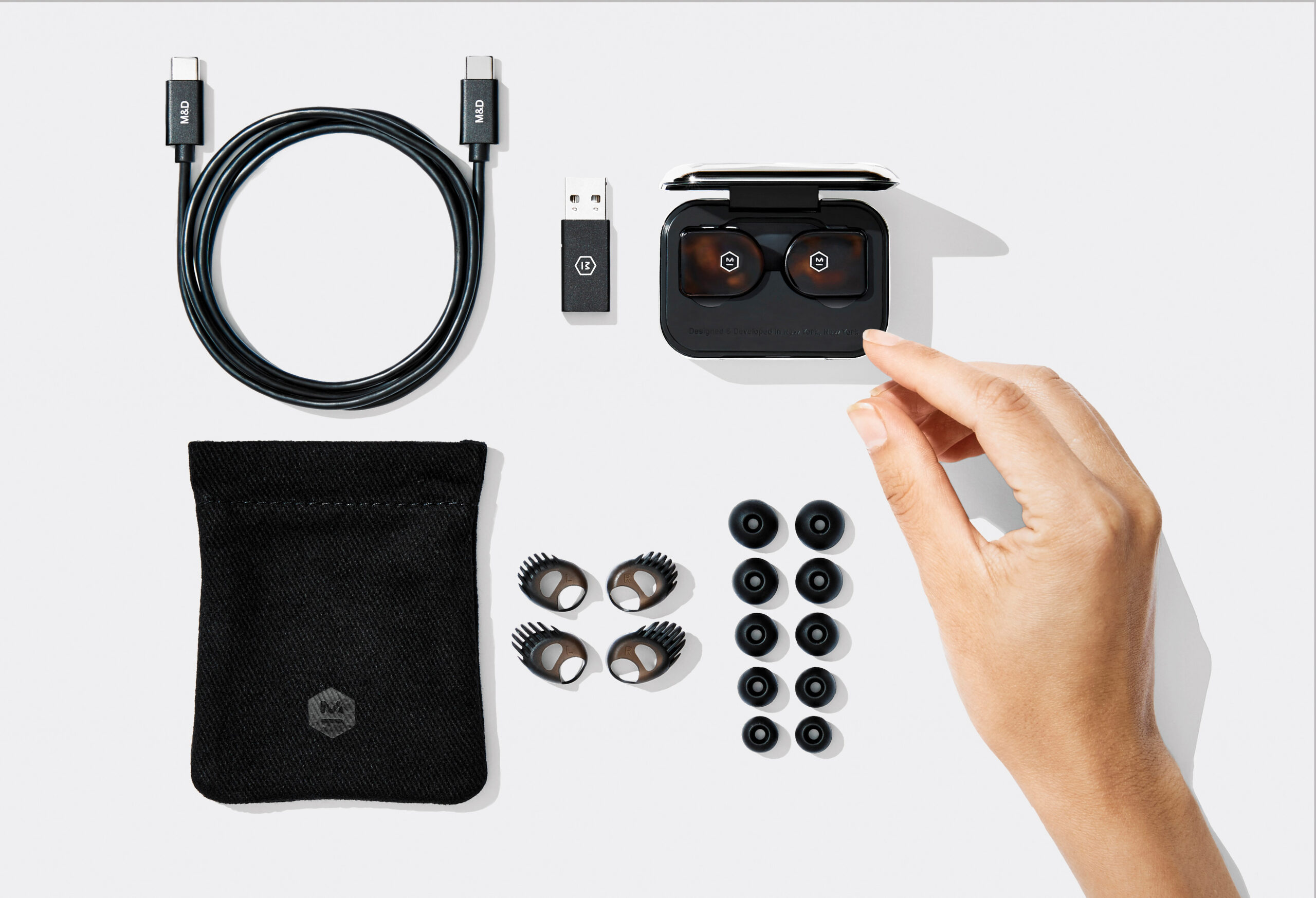 Product image of the Master &amp; Dynamic MW07 True Wireless. Image is overhead with all included accessories and a woman's hand reaching for the earbuds.
