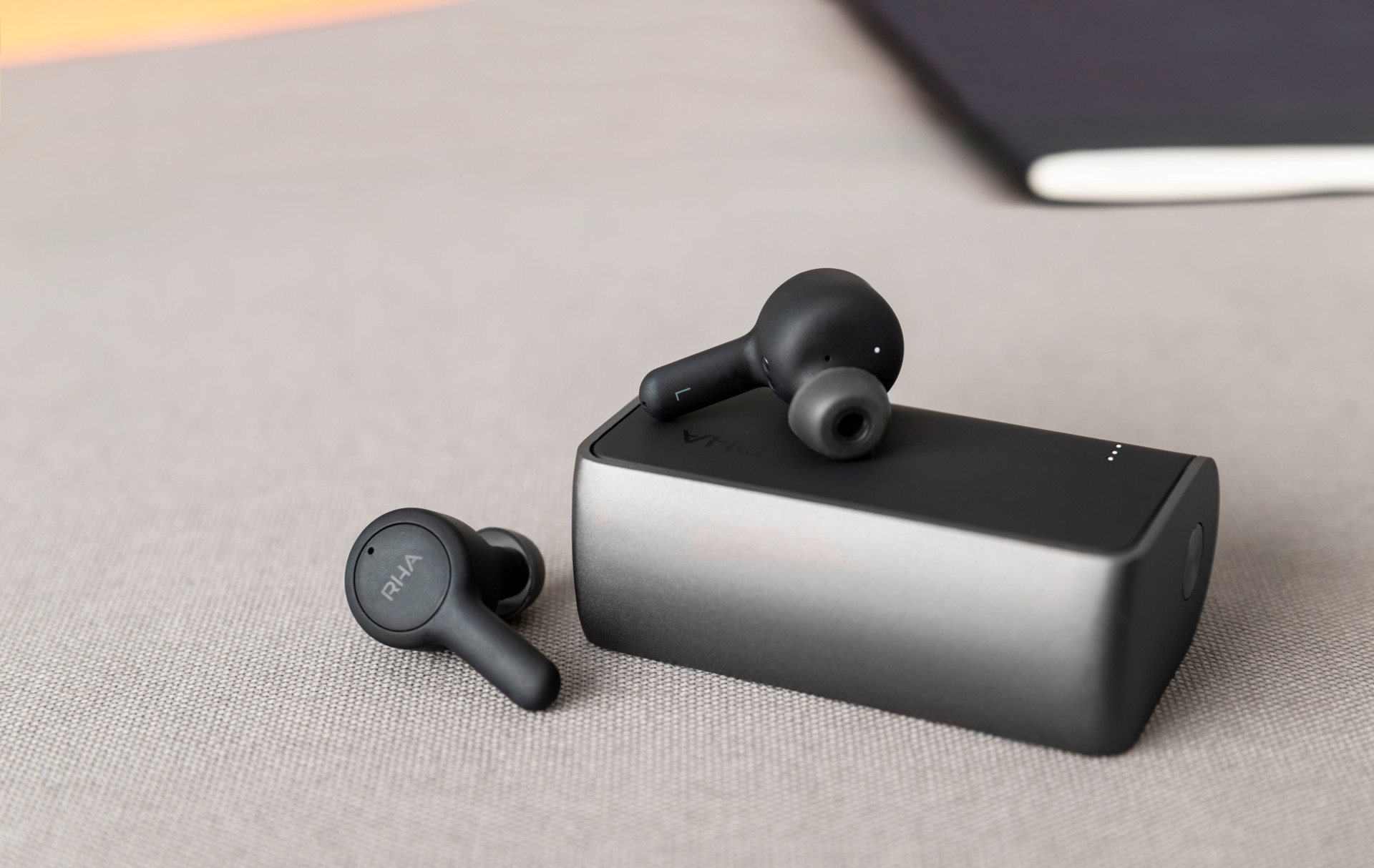 Product image of the RHA TrueConnect true wireless earbuds resting on the included charging case on beige surface.