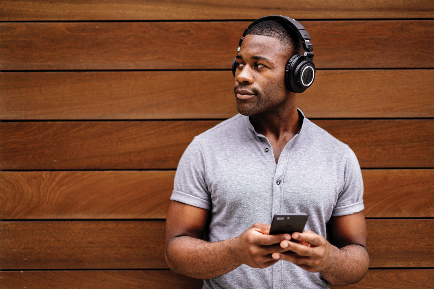 Audio-Technica ATH-M50xBT lifestyle image of man wearing headphones while he leans against a paneled wood wall.