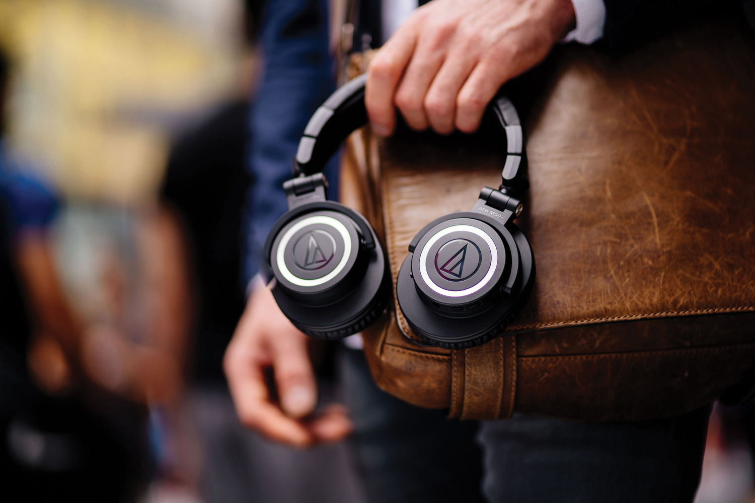 Audio-Technica ATH-M50xBT lifestyle image of the headphones being held by a man's hand against a leather messenger bag.