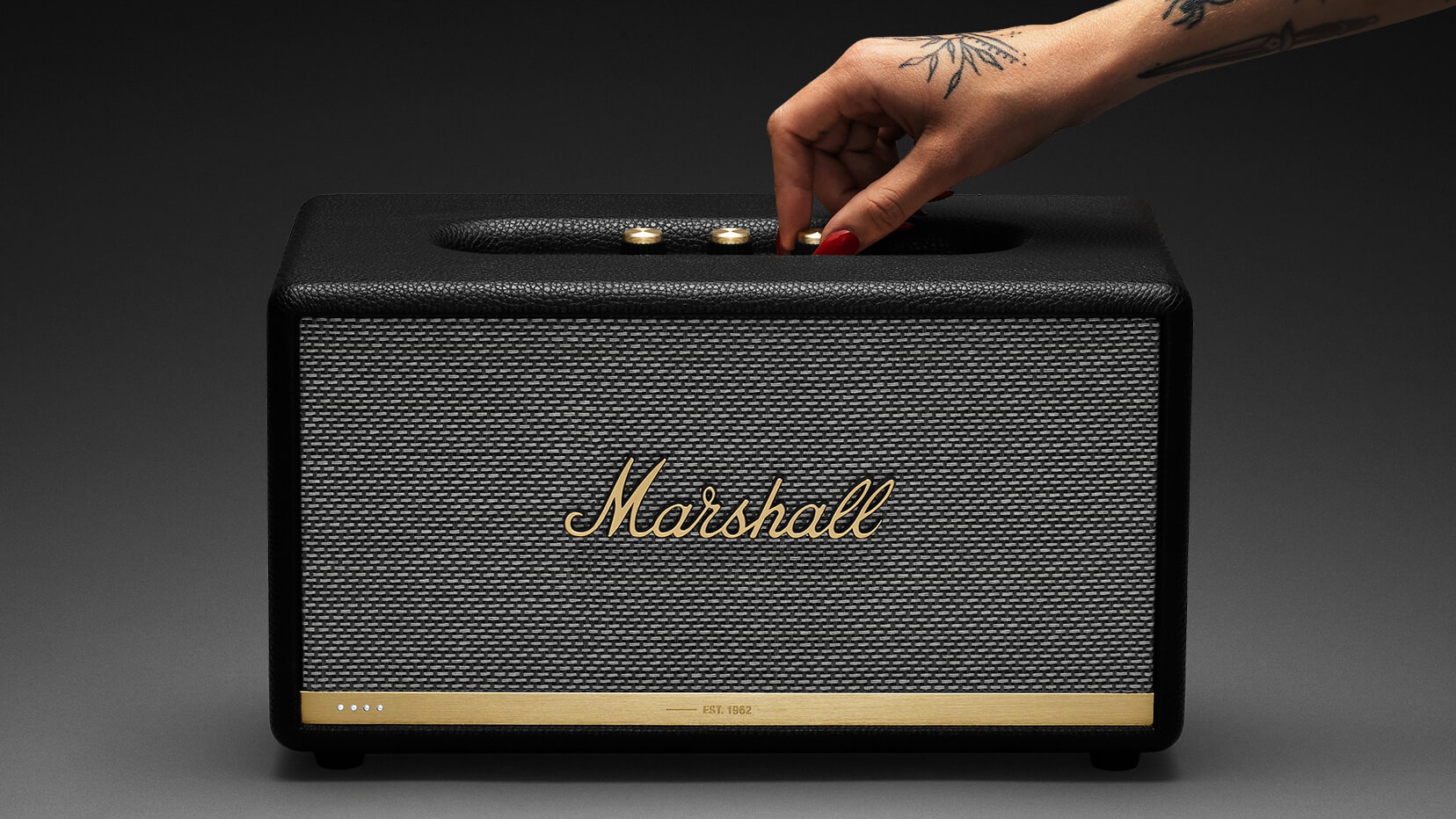 A product image of the Marshall Stanmore II Voice speaker against gradient background wtih a female hand turning one of the dials.