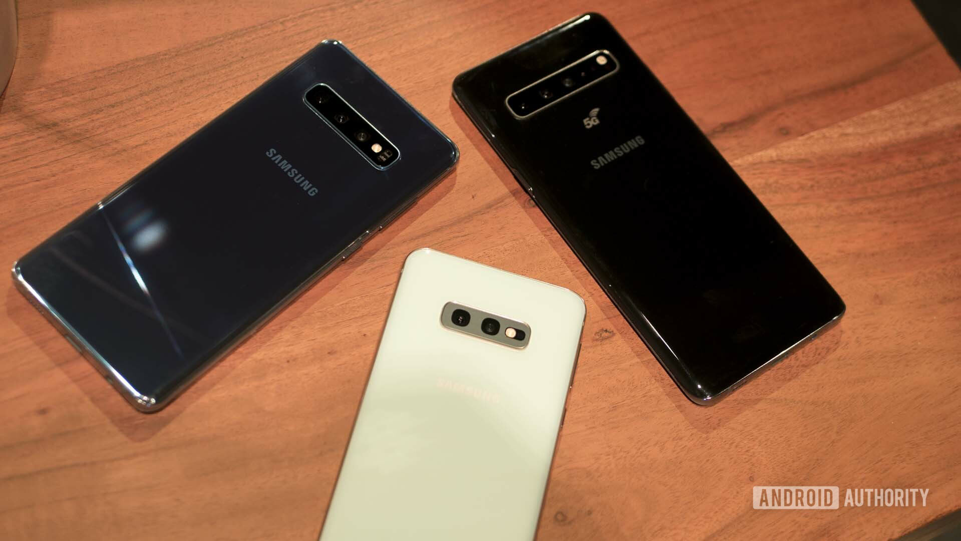 Back side photo of the Samsung Galaxy S10, Galaxy S10E and Galaxy S10 5G on a wooden table.