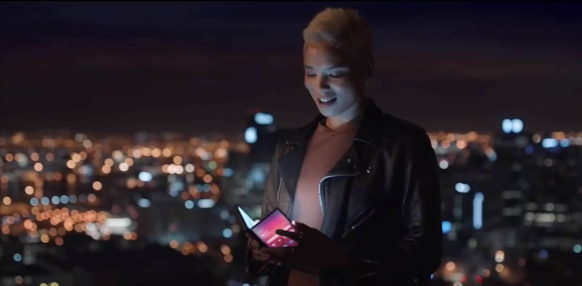 A woman holding a folding phone in front of a city at night.