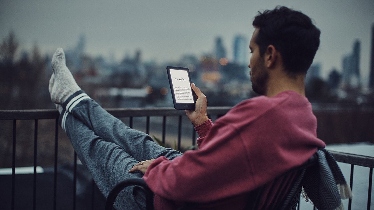 A promotional image of a man sitting on a balcony at night reading on an Amazon Kindle using its new front light.