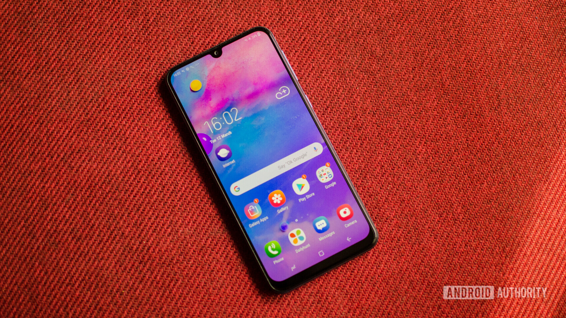 Front side of the Samsung Galaxy M30 laid on a red material, with the display turned on.