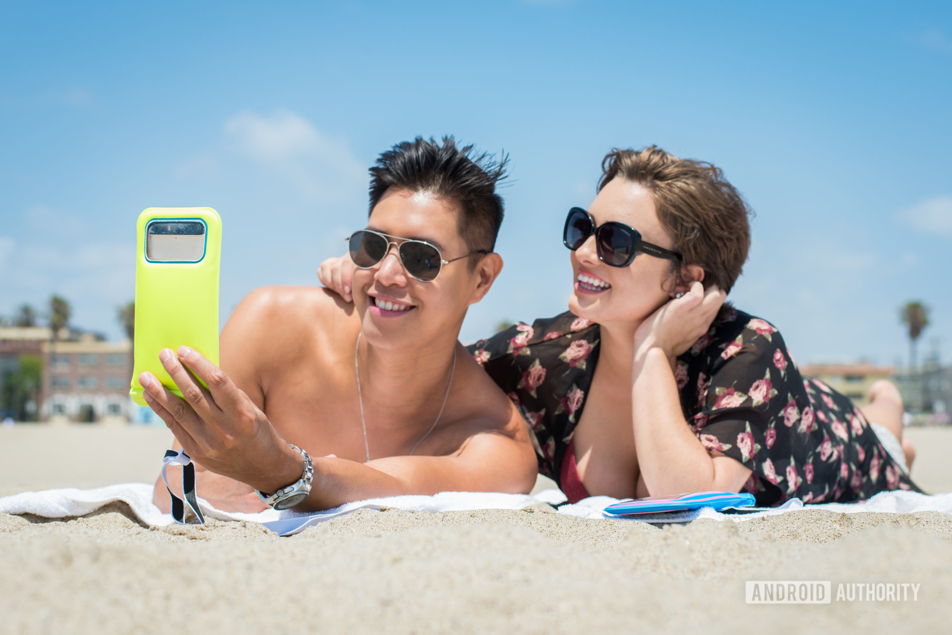 A man holding a yellow CaliCase waterproof pouch while a woman looks on next to him. They are both on the beach.