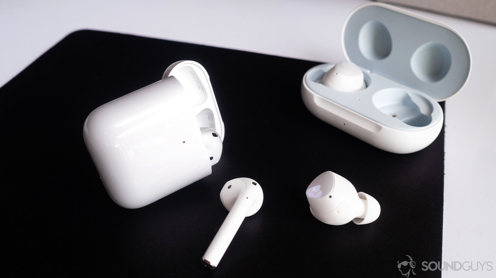 new AirPods (2019) and Samsung Galaxy Buds adjacent to one another on a table.