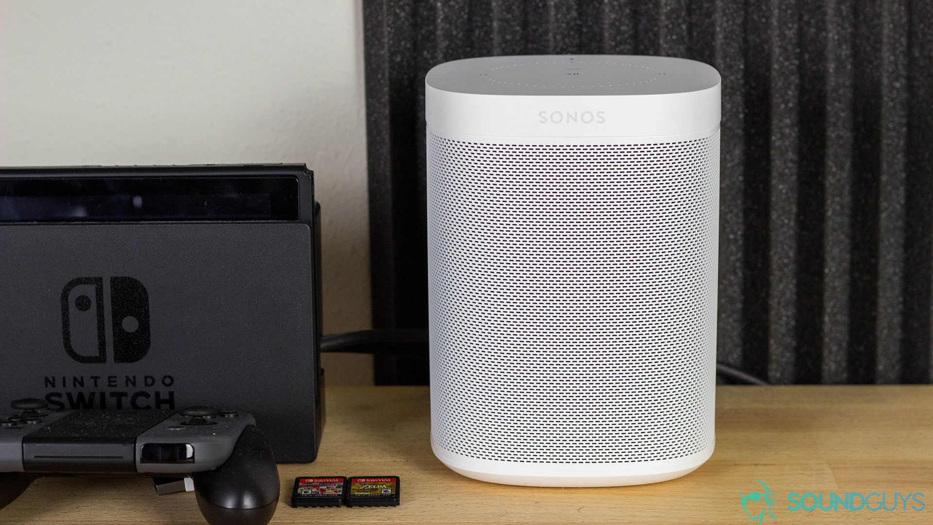 Sonos One in white next to a Nintendo Switch stand with game cartridges in front. The items are on a wood desk with soundproof foam in the background.