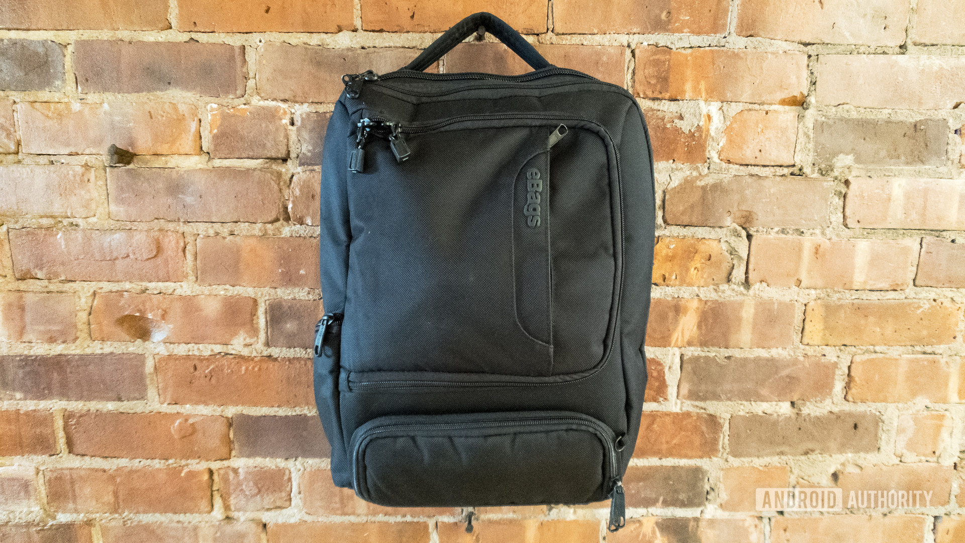 An image of the eBags Professional Slim Junior Laptop Backpack hanging on a brick wall.