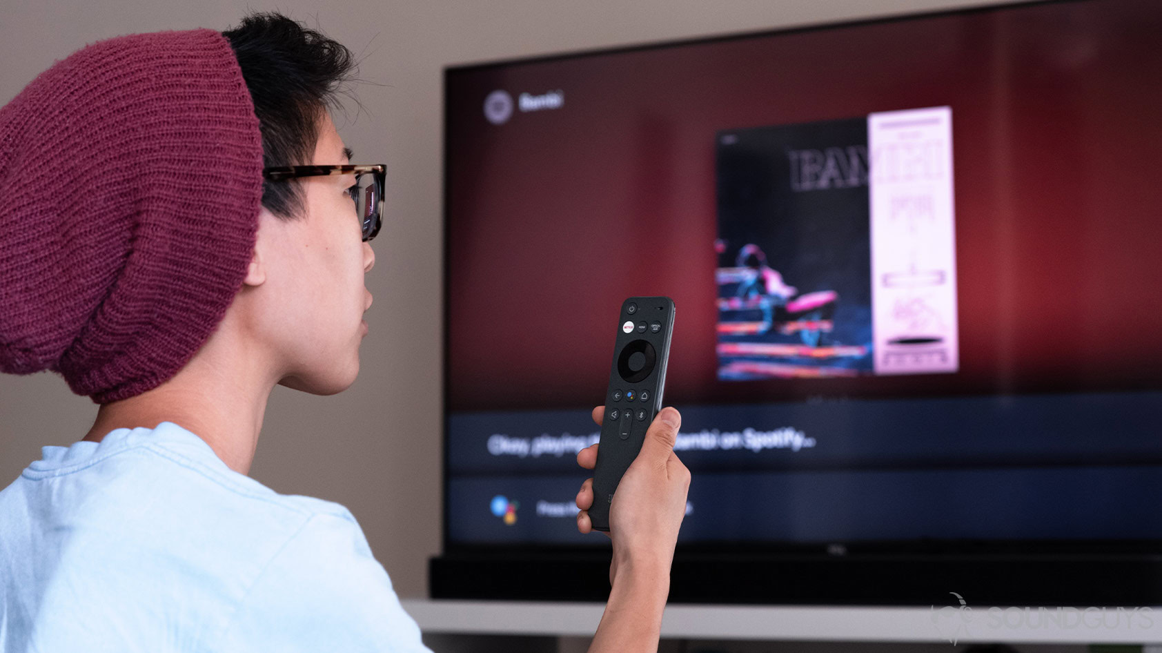 A woman holding the JBL Link Bar remote and speaking to Google Assistant to switch songs on Spotify.