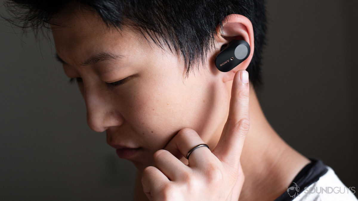 A woman wears the Sony WF-1000XM3 earbuds and reaching for the touch panel of the left 'bud.