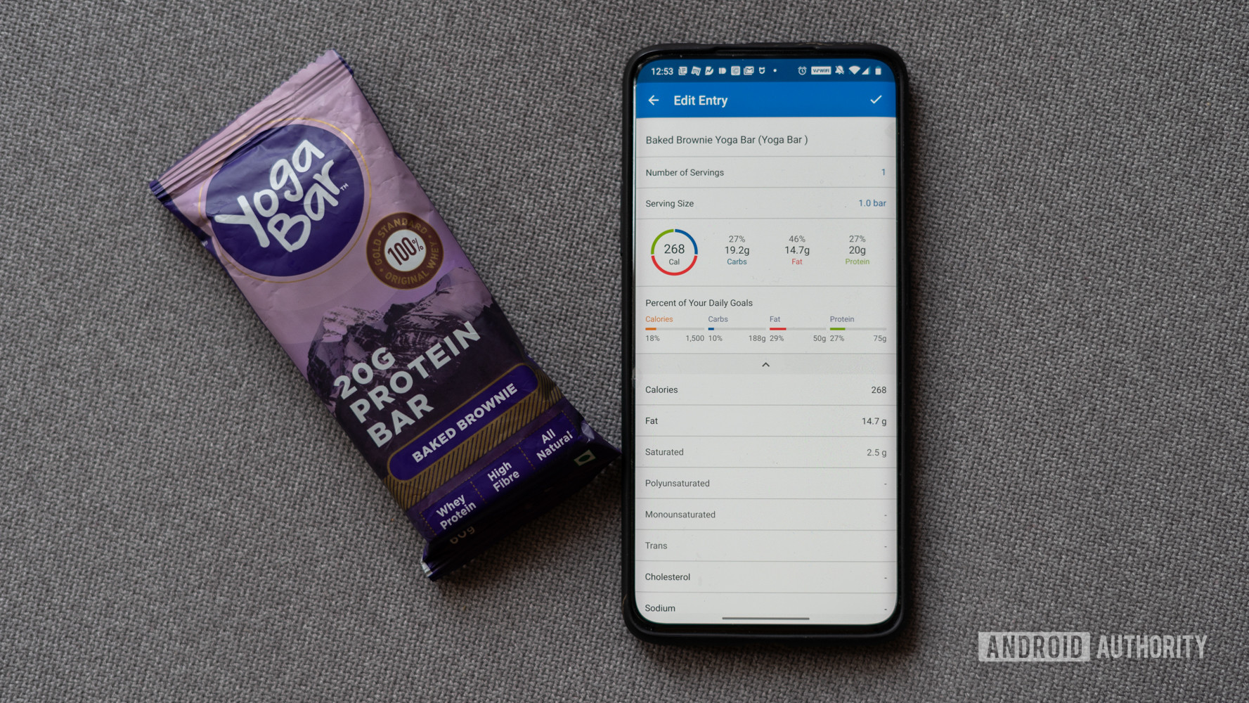 An iPhone displaying a user's calorie tracking data in the MyFitnessPal app rests on a linen surface next to a Yoga Bar protein bar.
