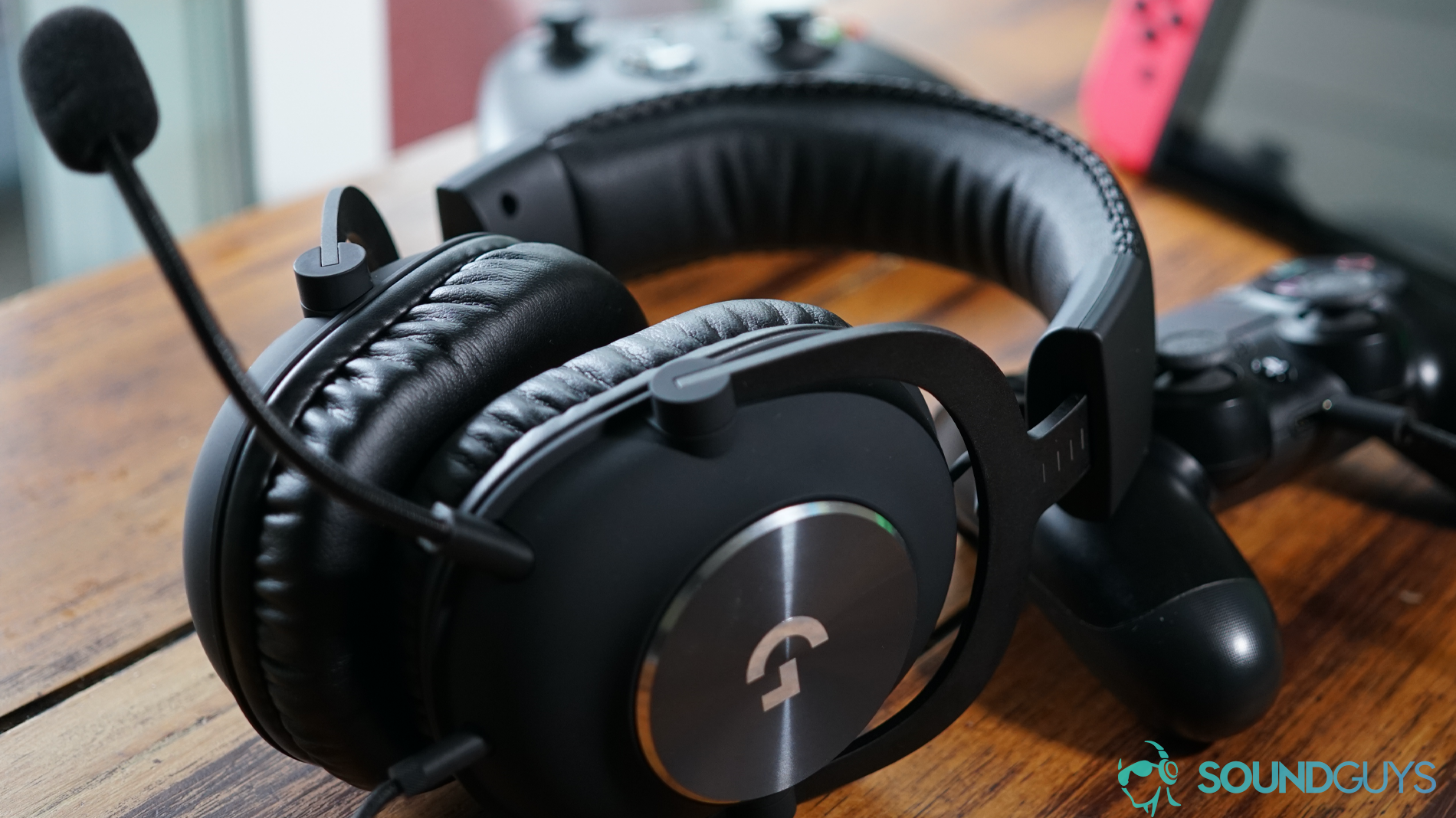 The Logitech G Pro X gaming headset sits on a wooden table in front of a Playstation 4 Dualshock controller, Xbox One controller, and a Nintendo Switch.