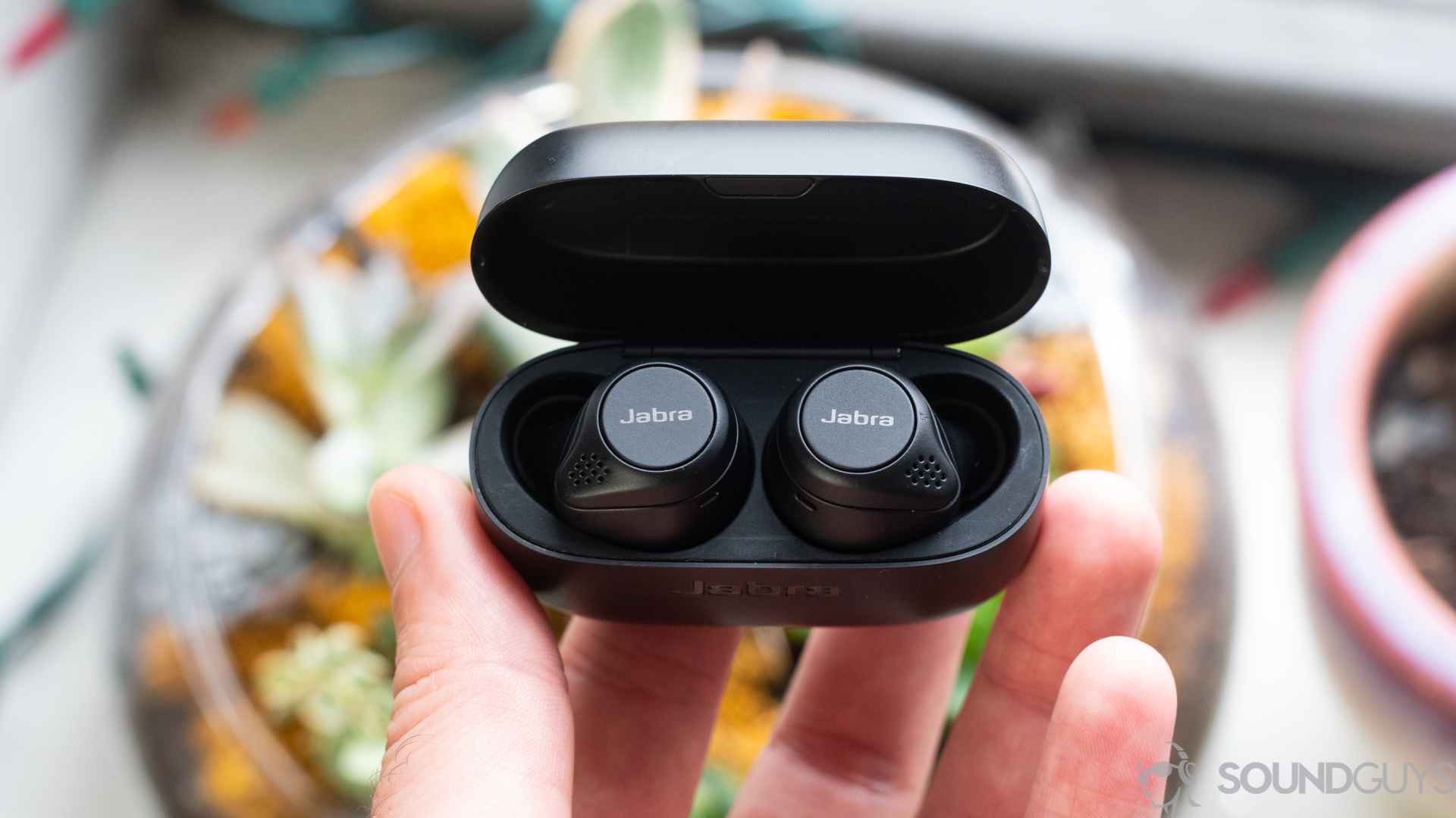 A picture of a man holding Jabra Elite 75t earbuds with case open over plants.