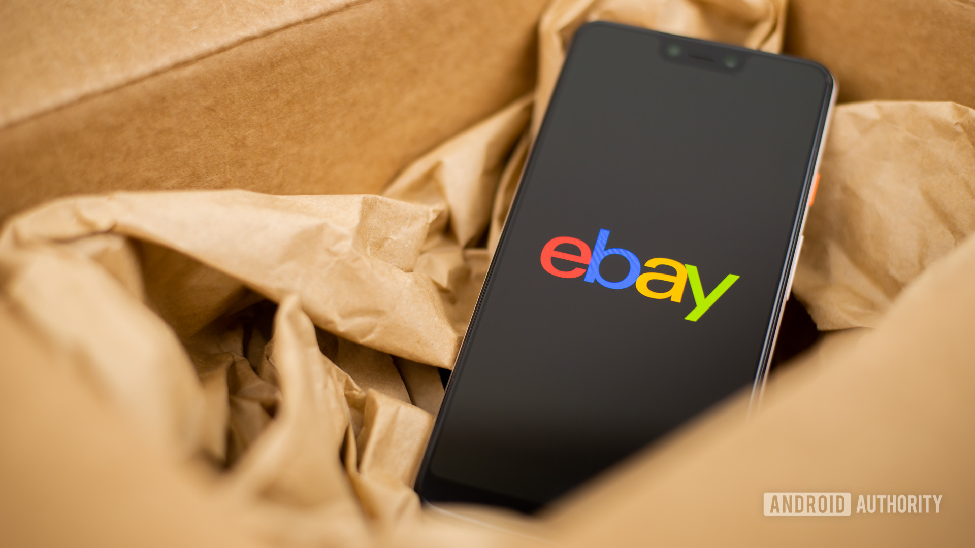 A smartphone sitting in brown paper in a brown cardboard box with a screen that reads &quot;ebay&quot; in red, blue, yellow, and green letters, respectively.