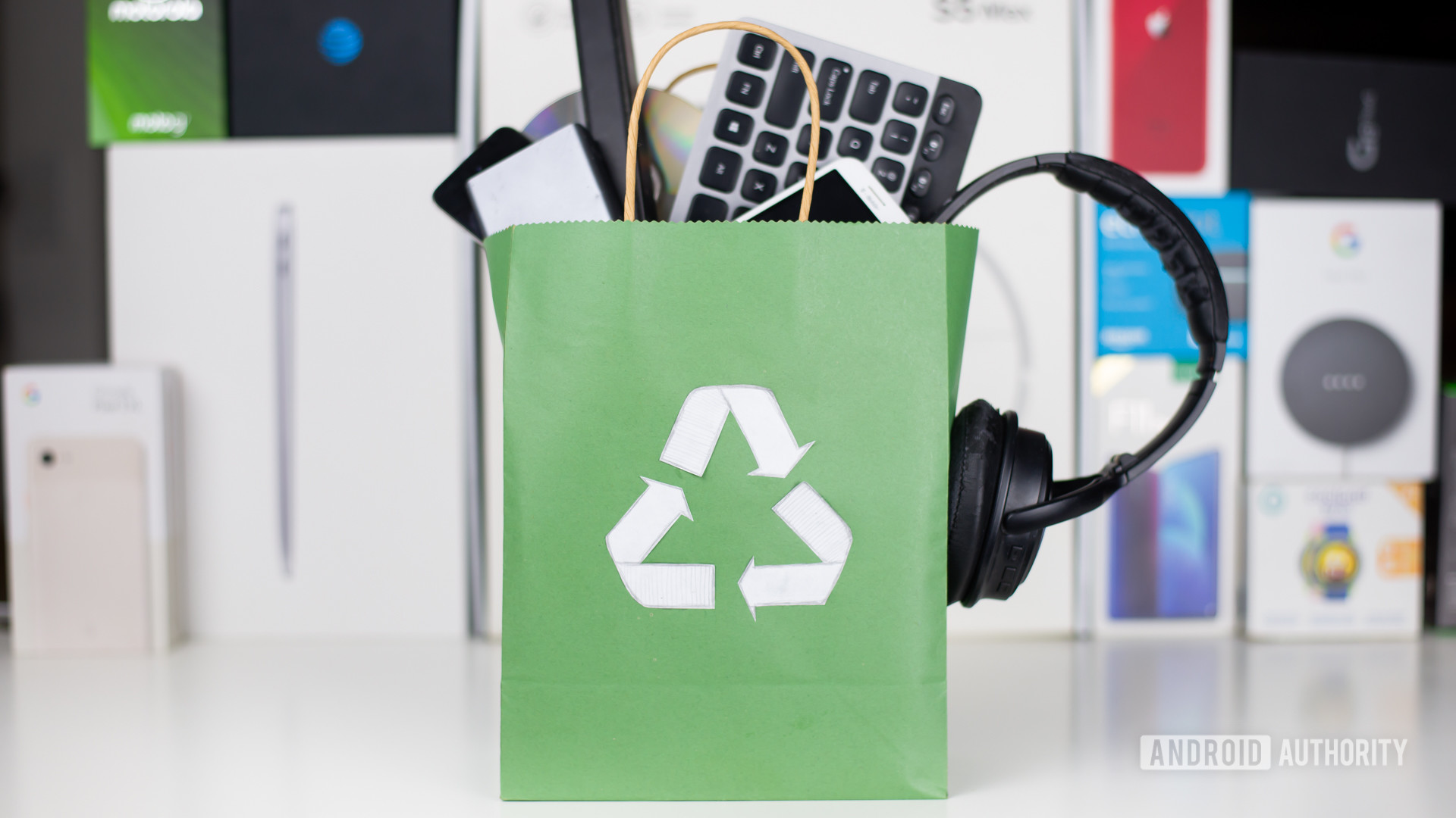 New electronics stuffed into a green paper bag with a white recyling symbol on it in front of electronic device boxes.