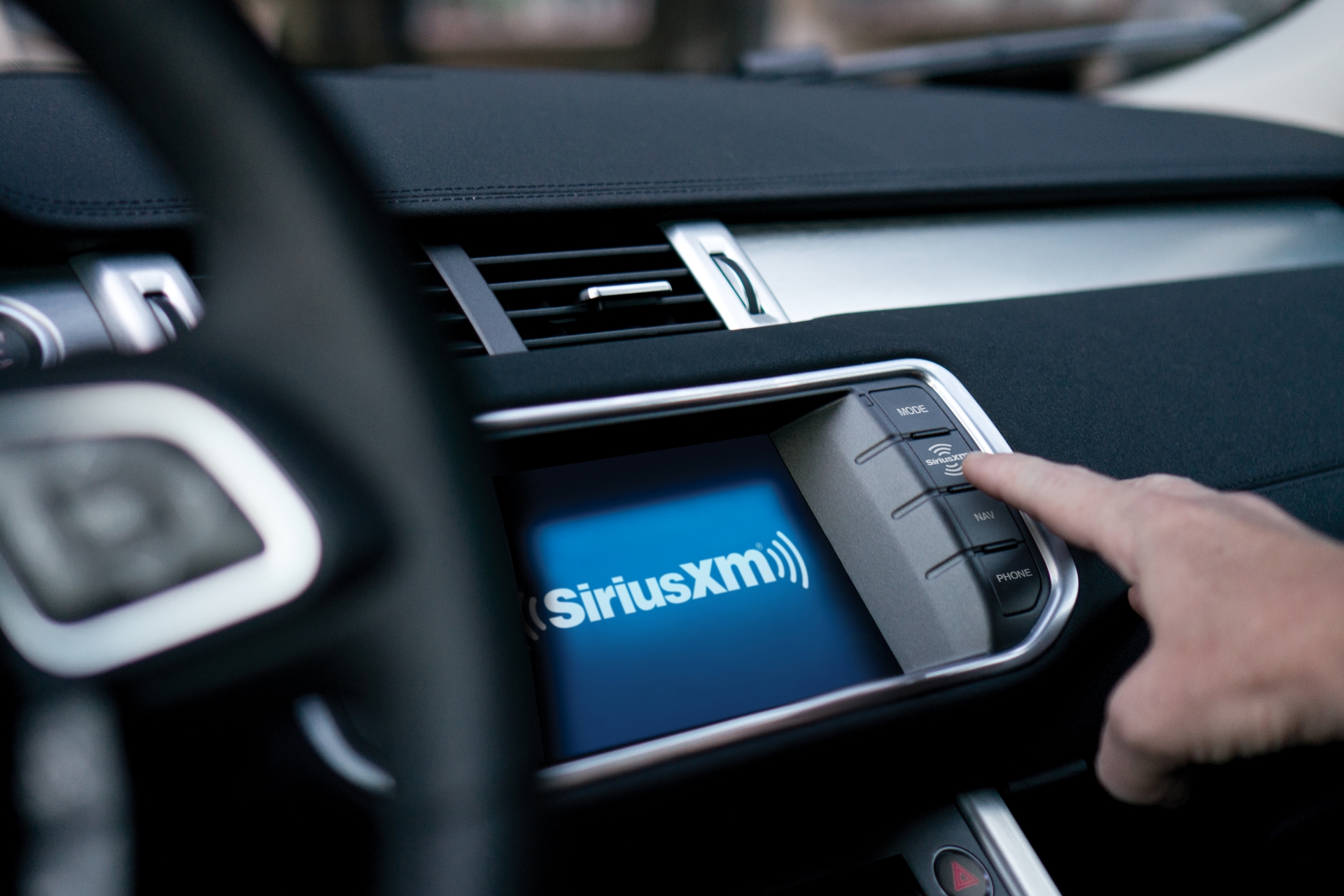 SiriusXM shown on the dashboard touch screen of a car with a finger pressing a button.
