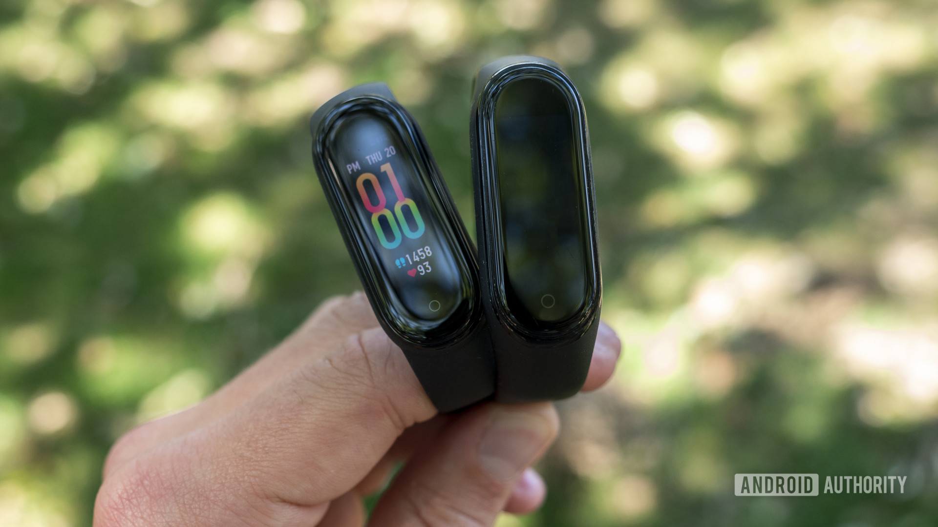 A user holds up a Xiaomi Mi Band 5 and a Xiaomi Mi Band 4 in front of greenery.