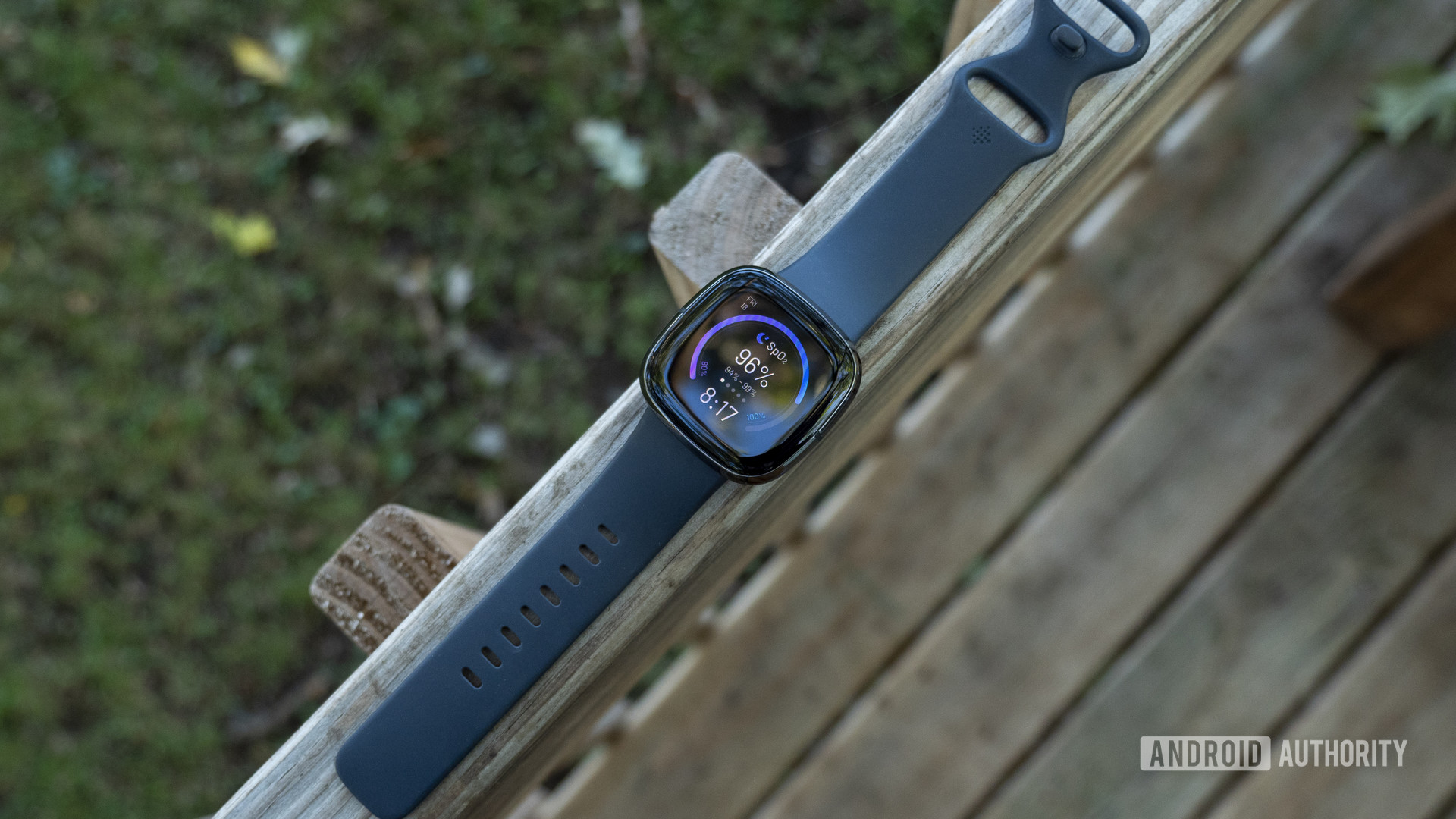 A Fitbit Sense rests on a wooden fence.