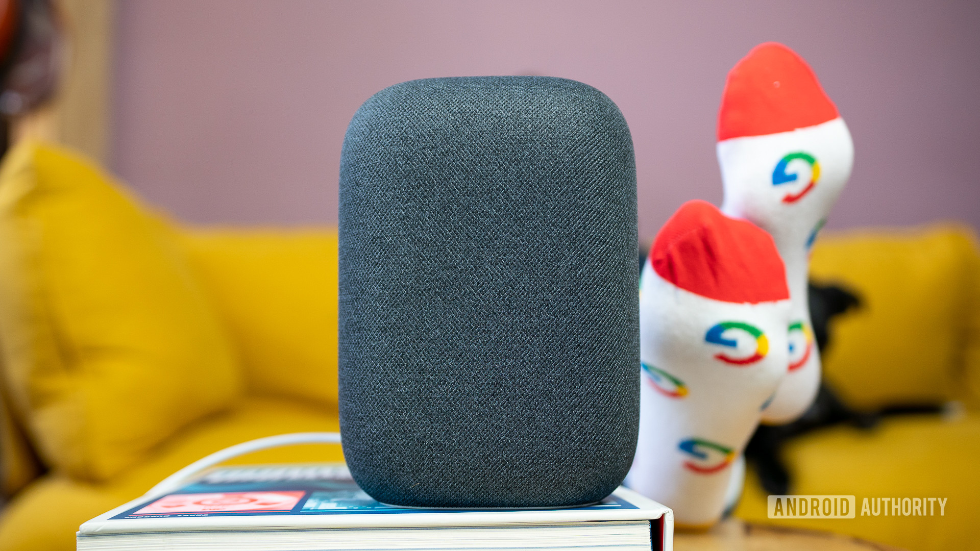 A gray Google Nest Audio on top of a book in front of a yellow sofa.