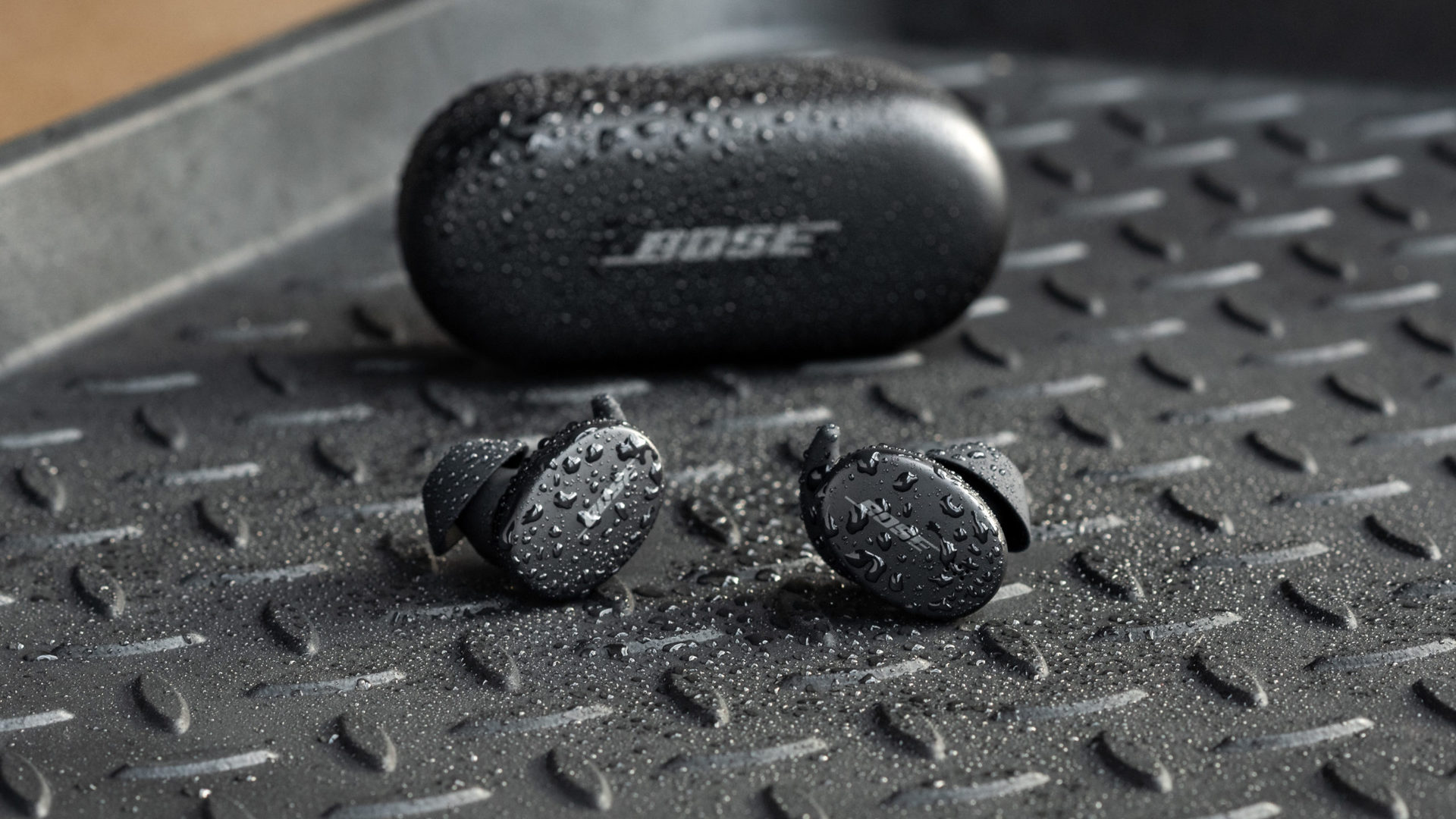 The Bose Sport Earbuds true wireless workout headphones are outside the sealed charging case, and all things are covered in splashes of water.