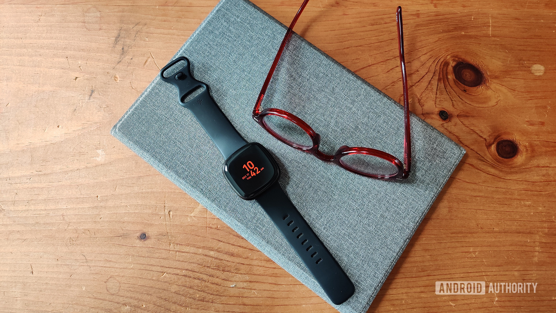 A Fitbit Versa 3 rests on a table alongside a pair of reading glasses.