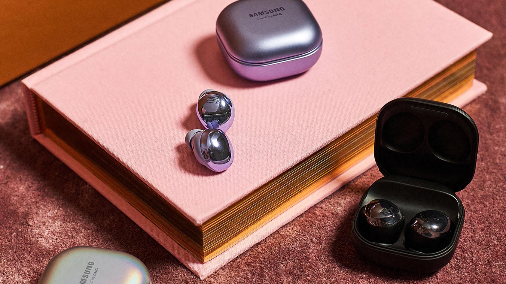 The Samsung Galaxy Buds Pro in purple, silver, and black, against a pink book and background.