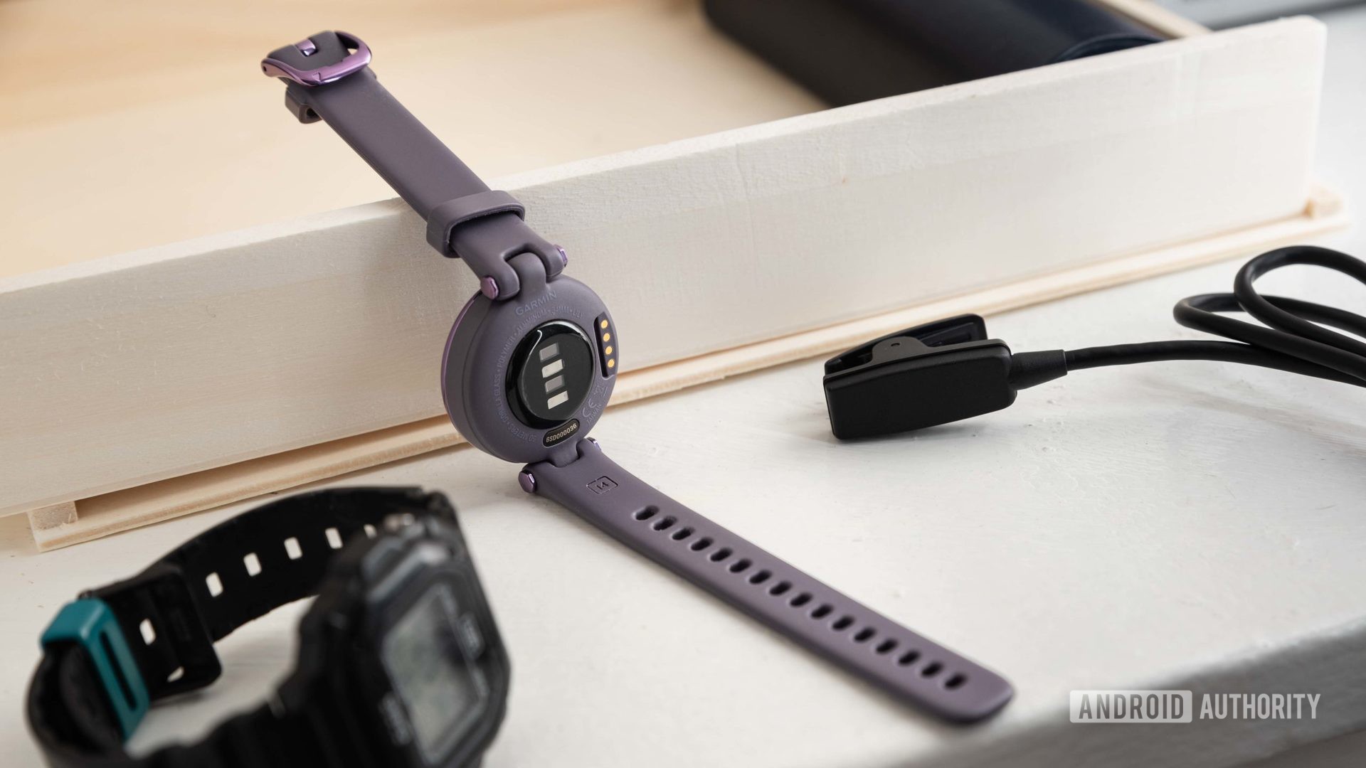 The Garmin Lily Sport Edition smart watch next to the proprietary charging cable and a Casio watch.