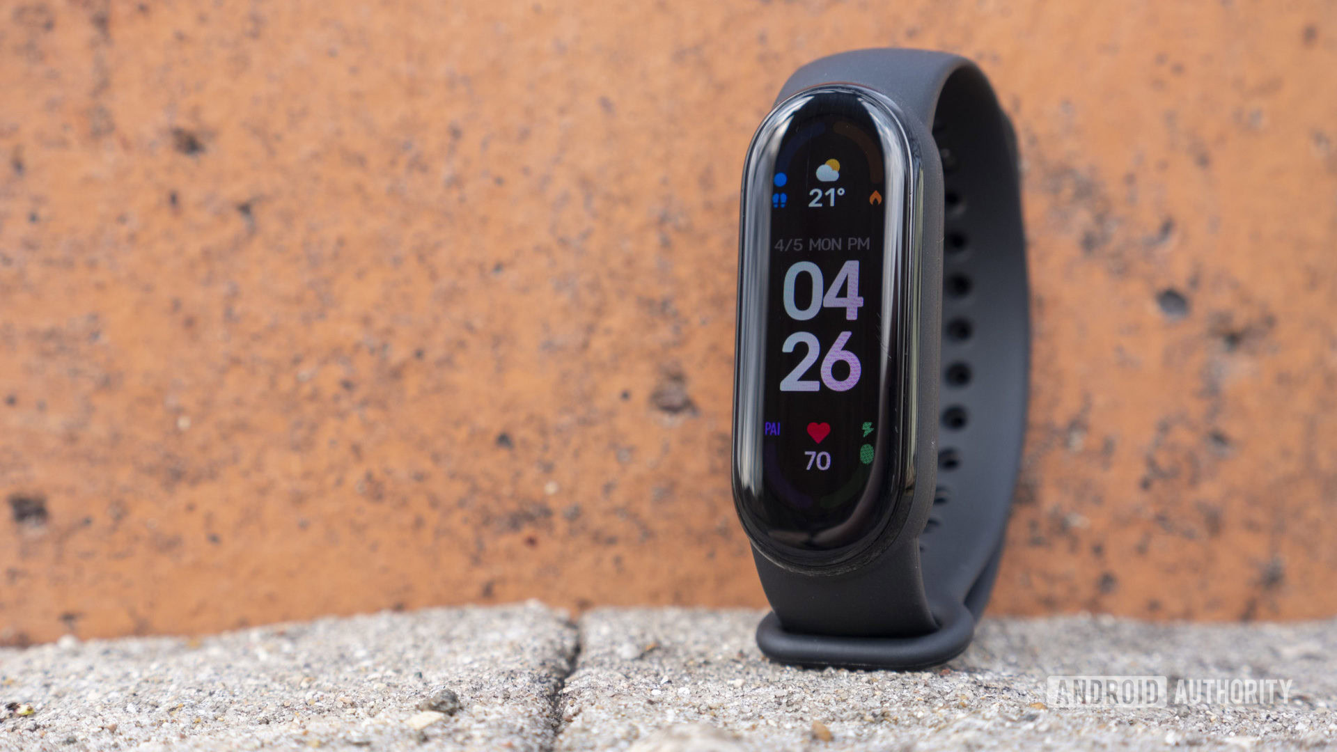 A Xiaomi Mi Band 6 is perched on a cement surface in front of an orange cement wall.