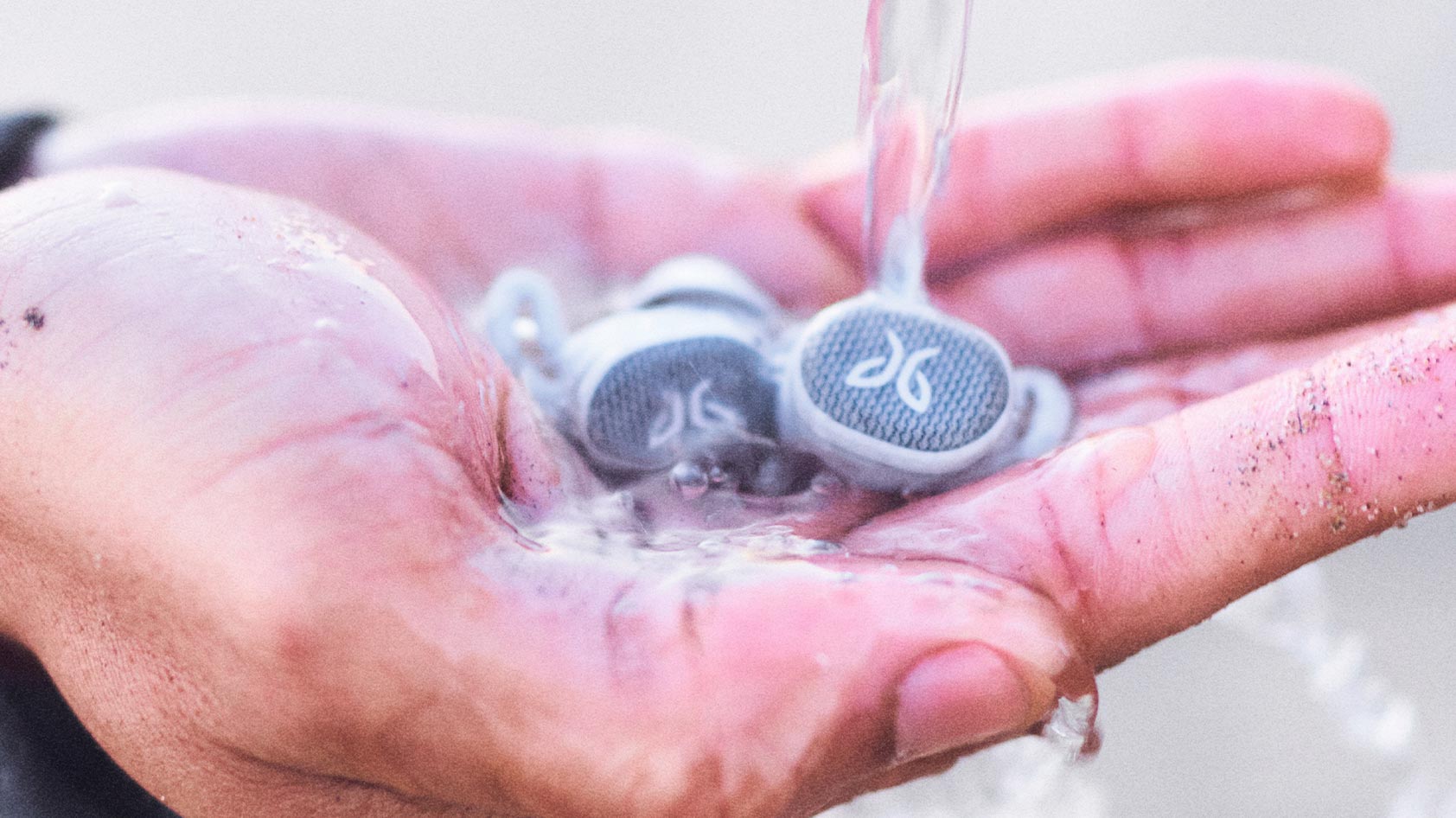 A hand holds the Jaybird Vista 2 true wireless workout earbuds as a stream of water pours down onto them.