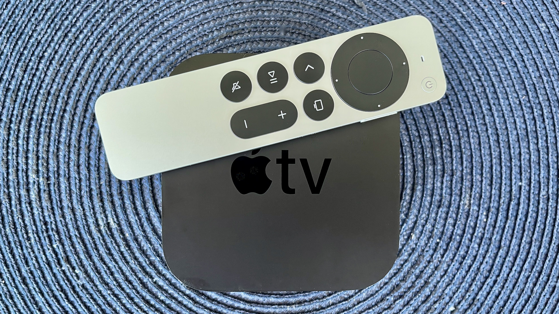 An Apple TV 4K with its remote on top of it lying on a carpet made of blue yarn woven into a circle.