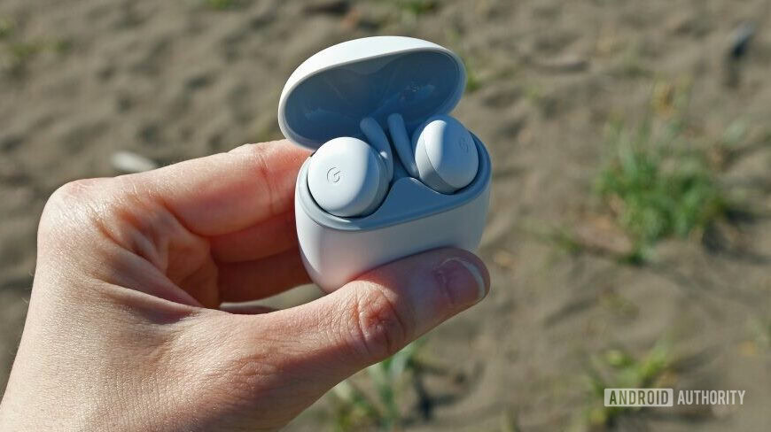 A hand holds the open case of the Google Pixel Buds A-Series in front of a beach.