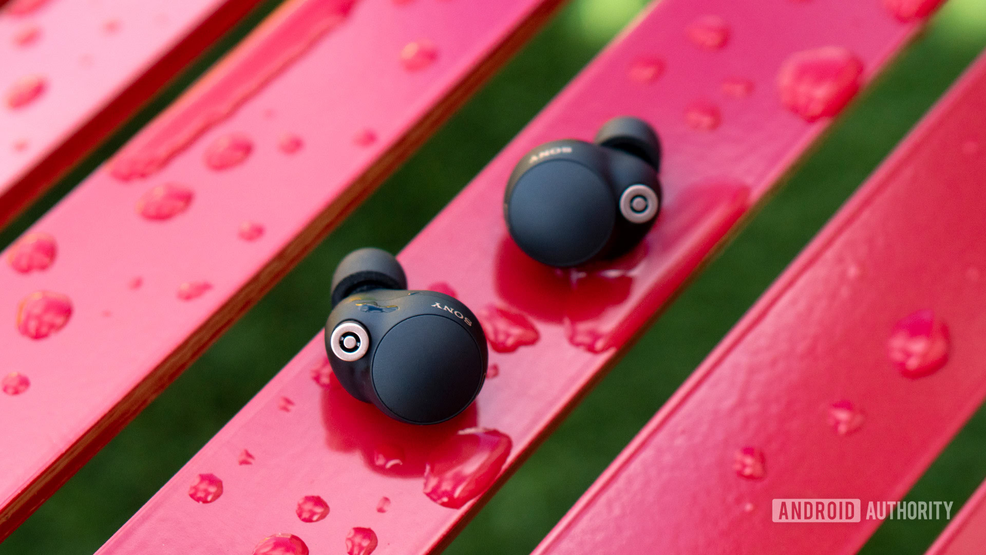 Sony WF-1000XM4 earbuds sitting outside of their case on a red bench covered with water droplets.