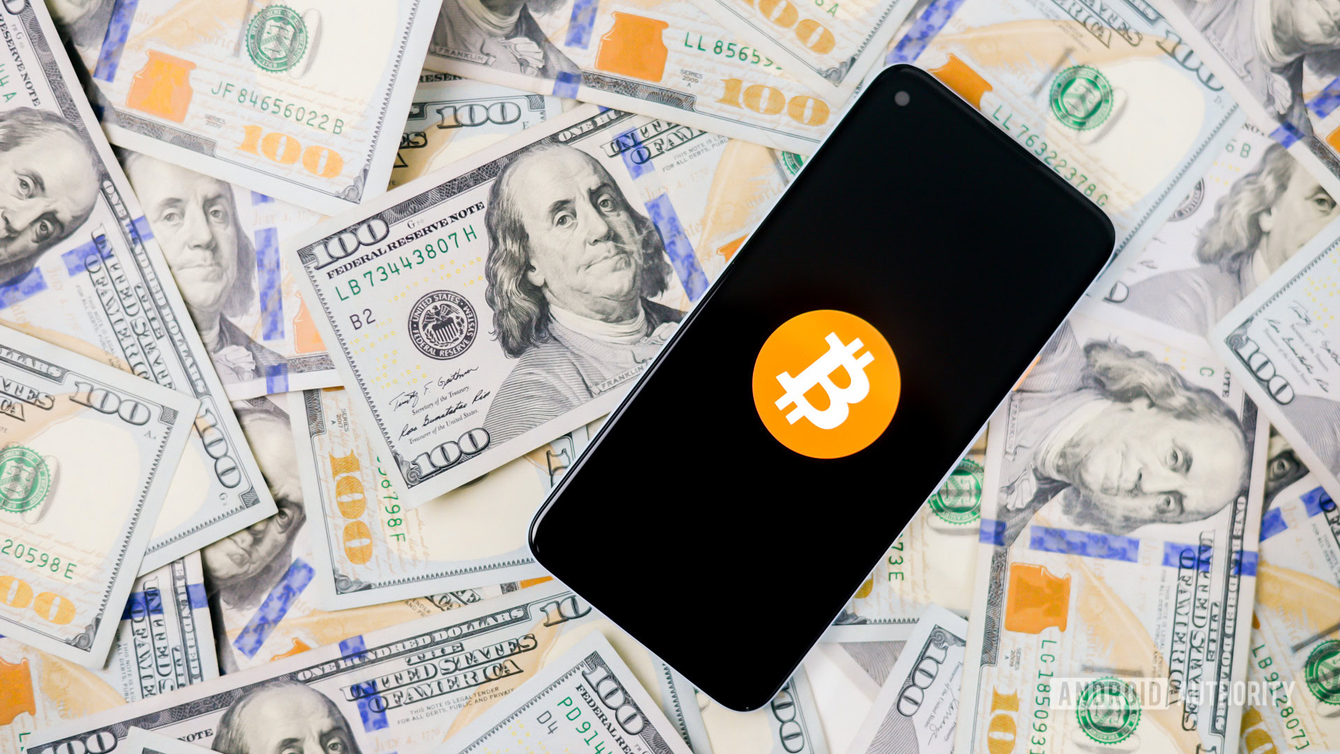 Stock image of the Bitcoin logo on a darkened smartphone screen resting on top of a stack of $100 bills.