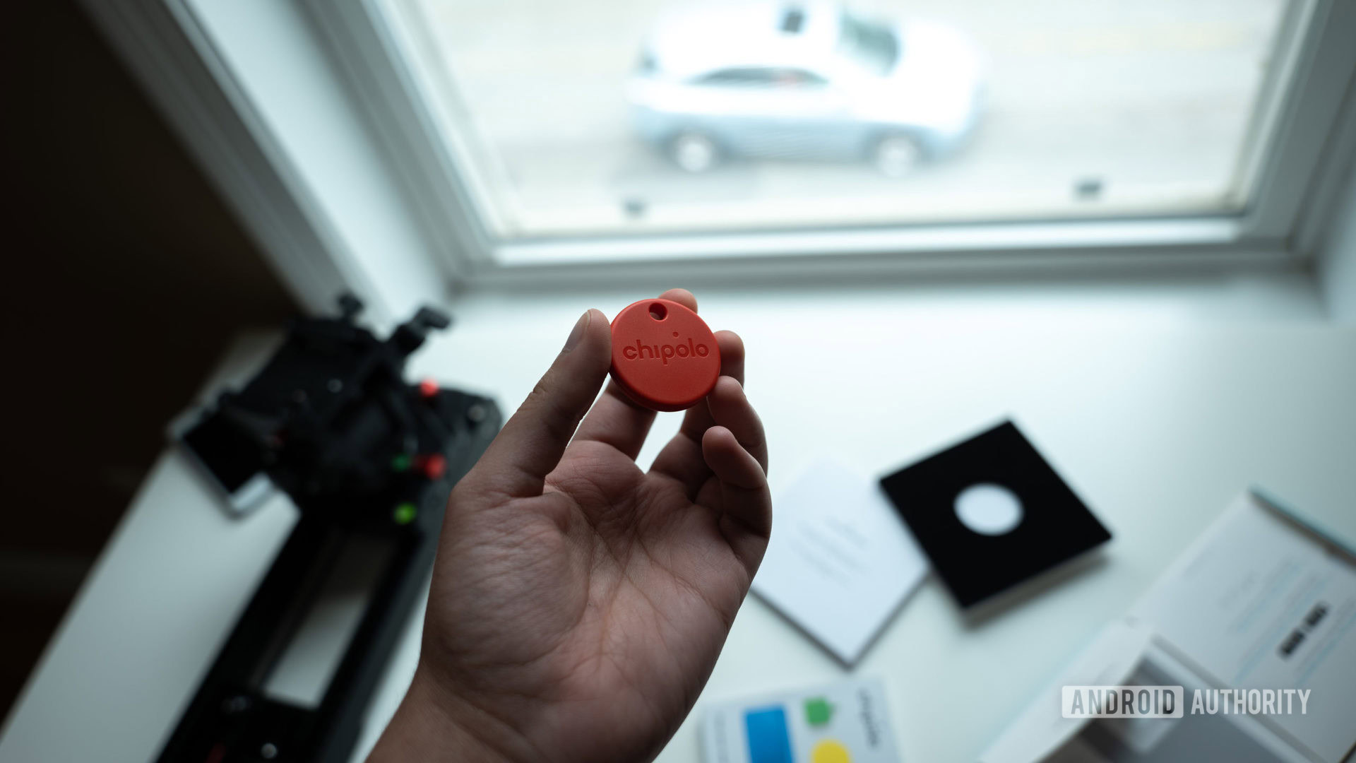 A picture of the Chipolo One tracker held above its packaging in front of a window