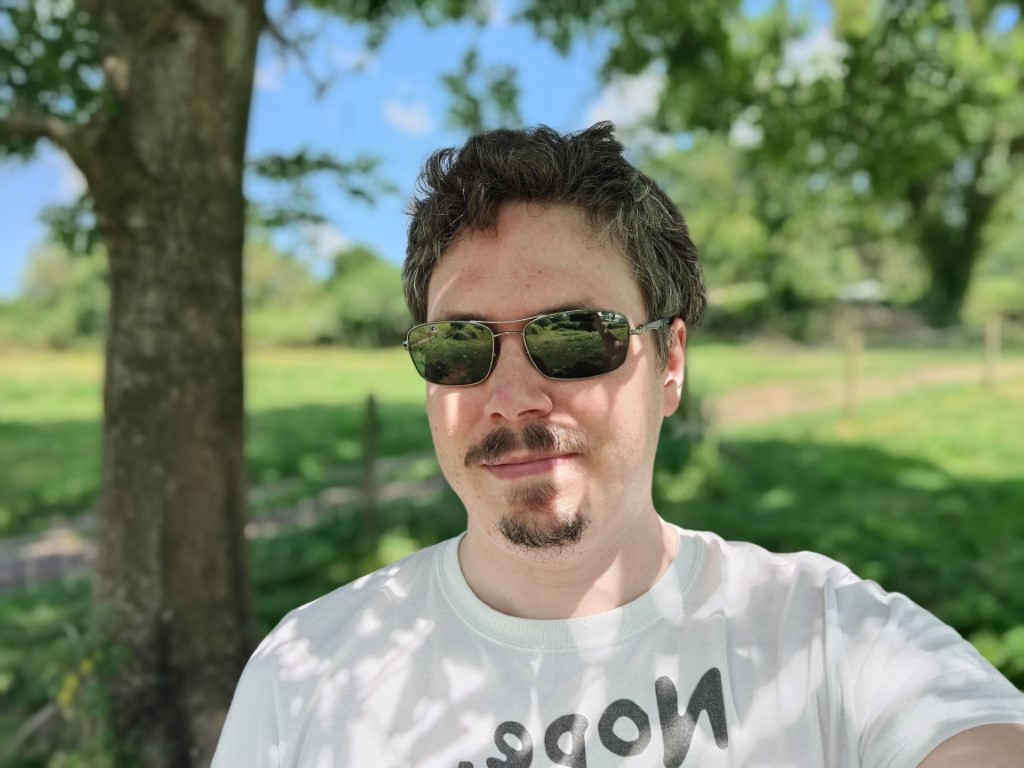 OnePlus 9 camera selfie 1 bokeh of a man in a white t-shirt wearing shades, with brown hair and a brown beard.