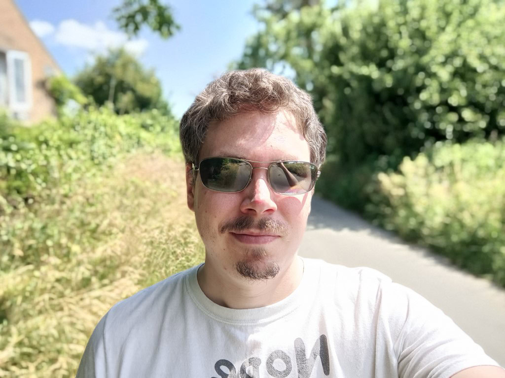 OnePlus Nord 2 5G camera sample selfie outdoors with bokeh showing a man in a light colored tshirt with brown hair and a beard, wearing shades.