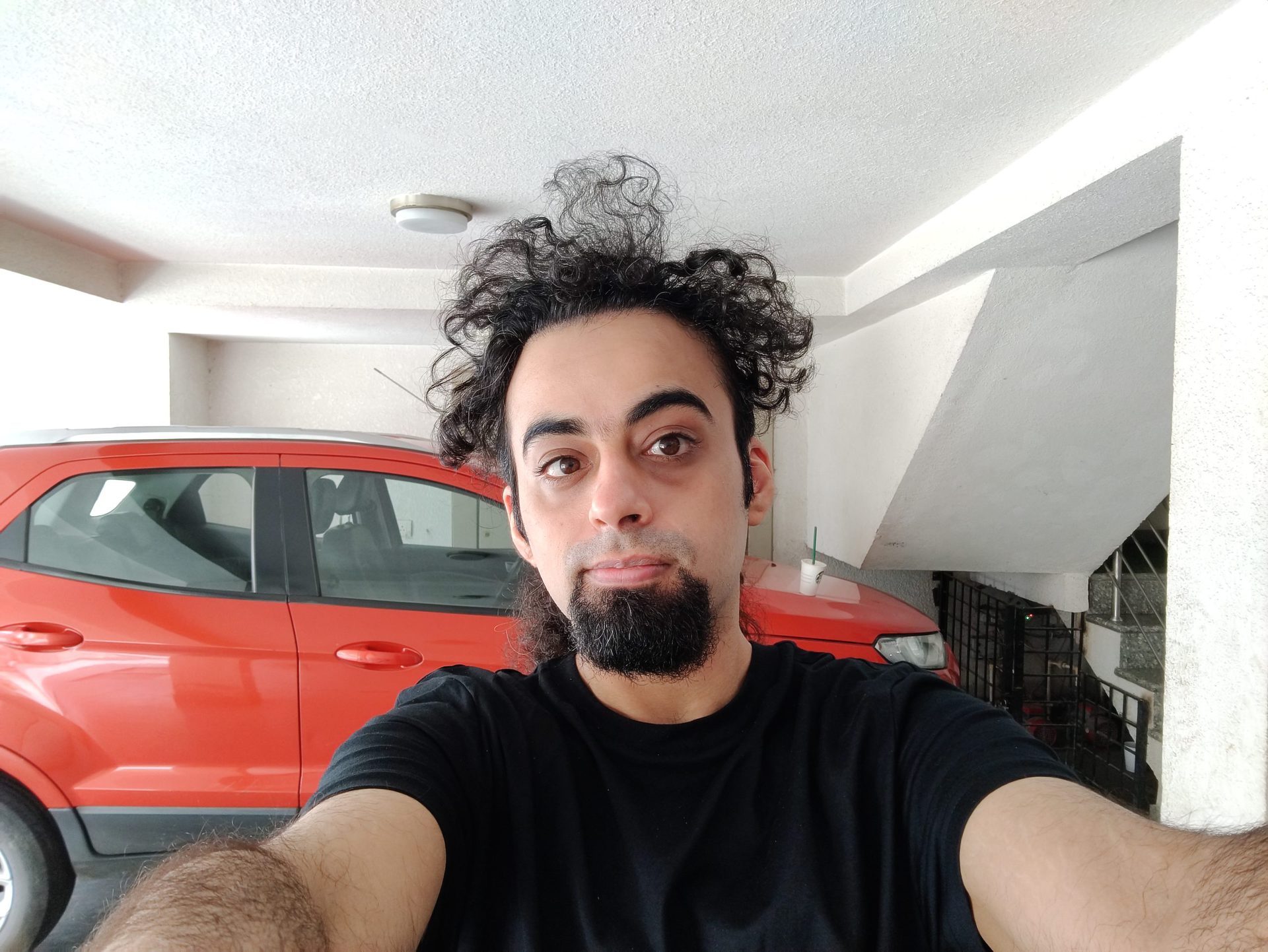The OnePlus Nord primary selfie indoors of a man with black hair and a beard, wearing a black t-shirt and standing in front of a red vehicle.