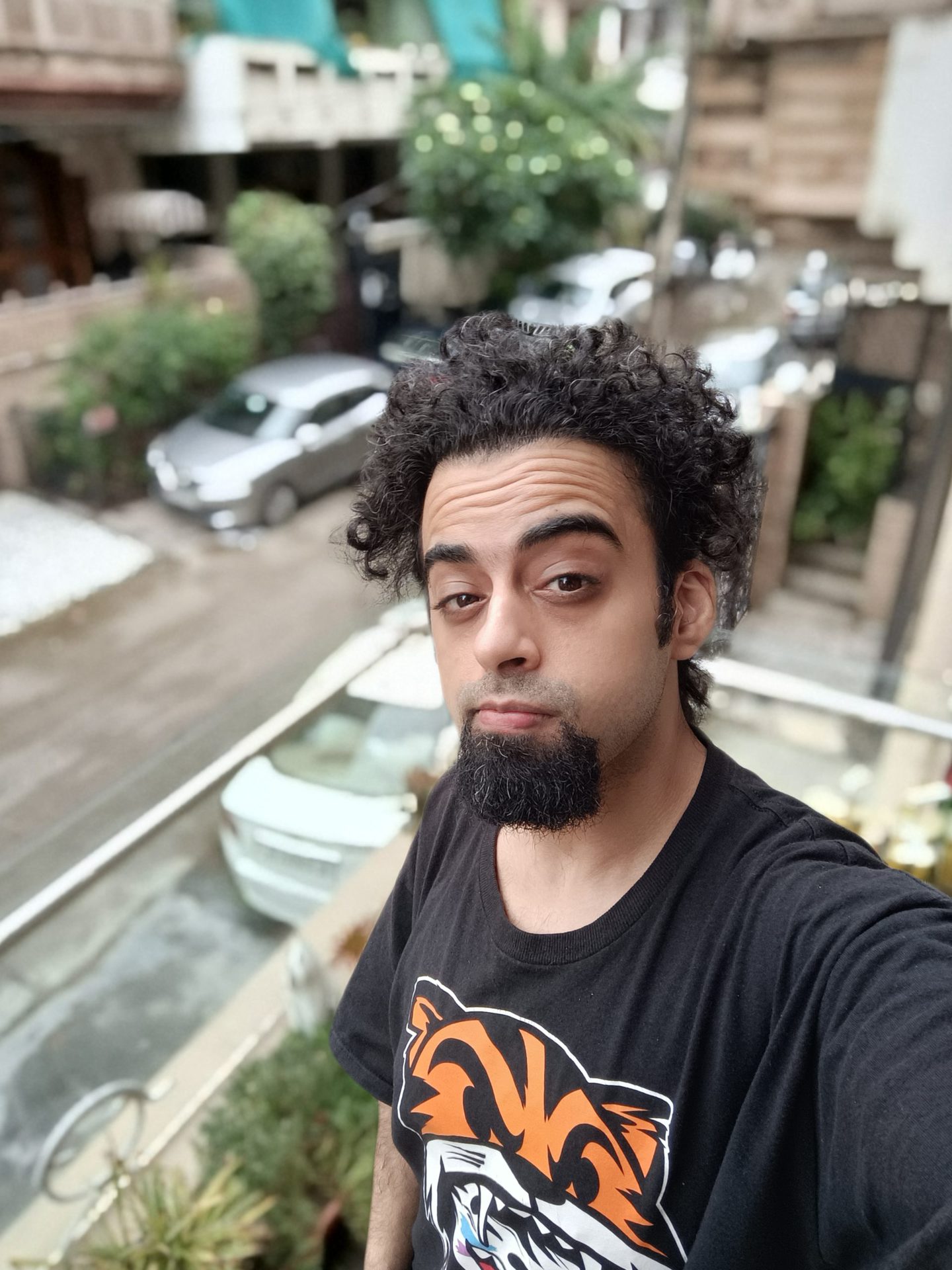 The Oppo Reno 6 Pro selfie portrait mode showing a man with black hair and beard in a black t-shirt with orange pattern standing on a balcony above the street below.