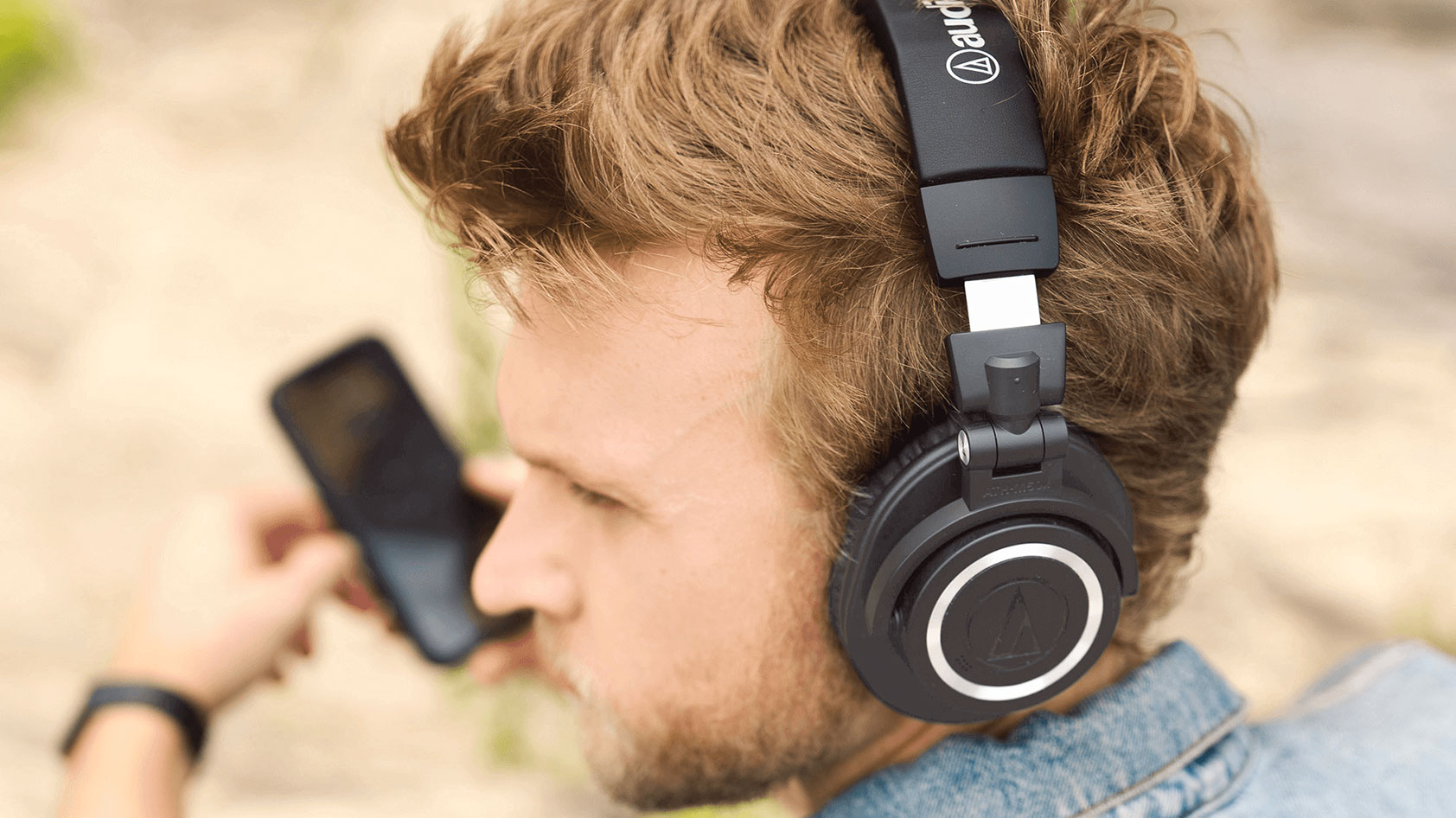A man wears Audio Technica at m50xbt2 on the beach holding a smartphone.