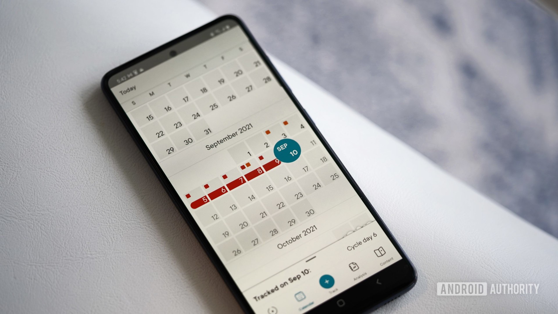 A Galaxy A51, open to the Clue app tracking calendar, rests on a white leather chair.