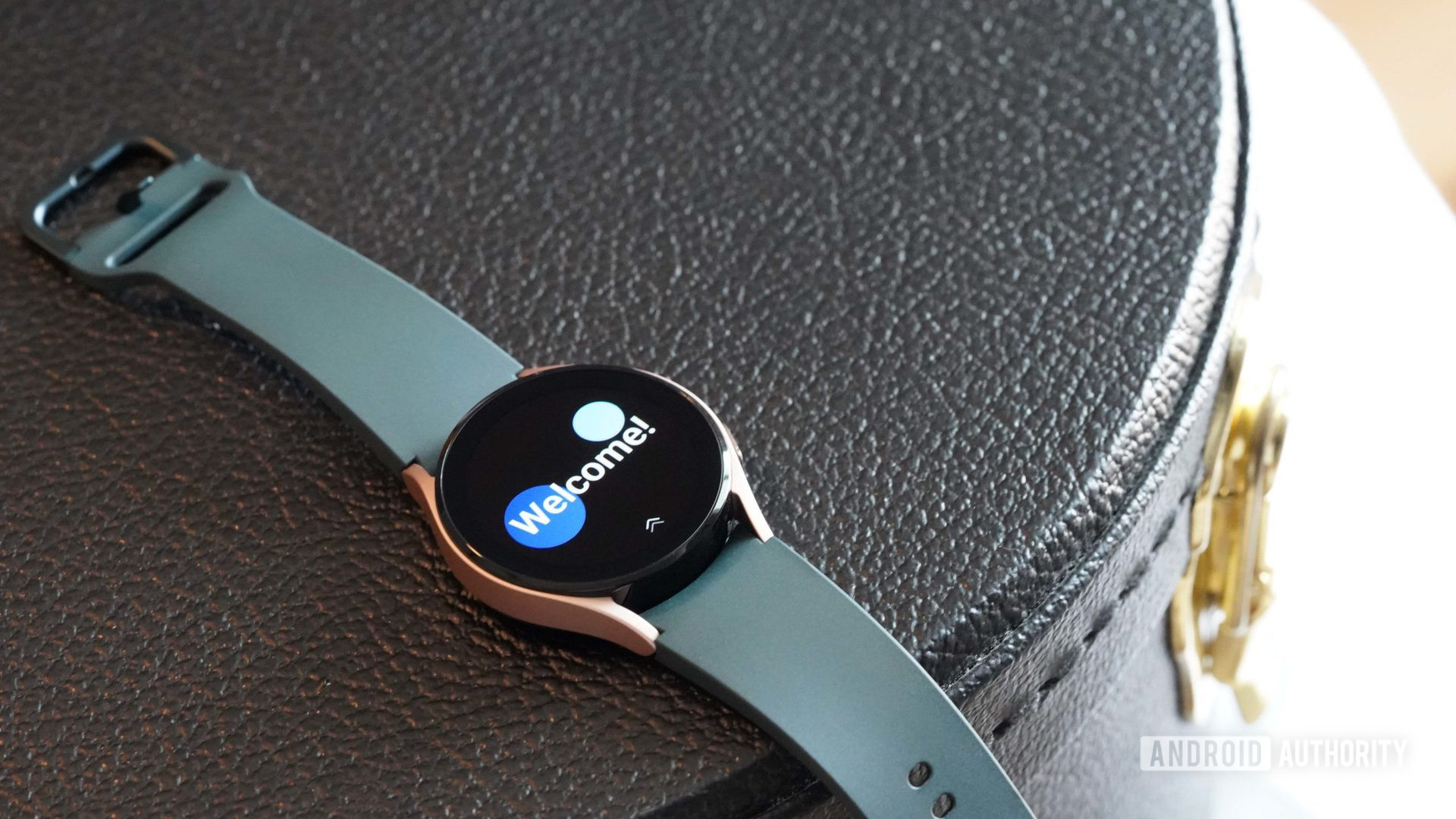 Bir Samsung Galaxy Watch 4, saatlerin Karşılama ekranını gösteren siyah bir deri çantanın üzerinde duruyor.