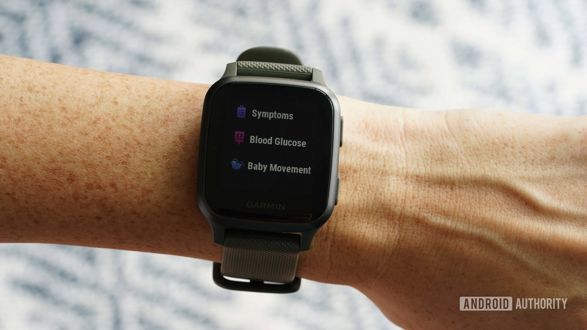 A Garmin Venu Sq on a woman's wrist displays the symptom, blood glucose, and baby movement logging screen.