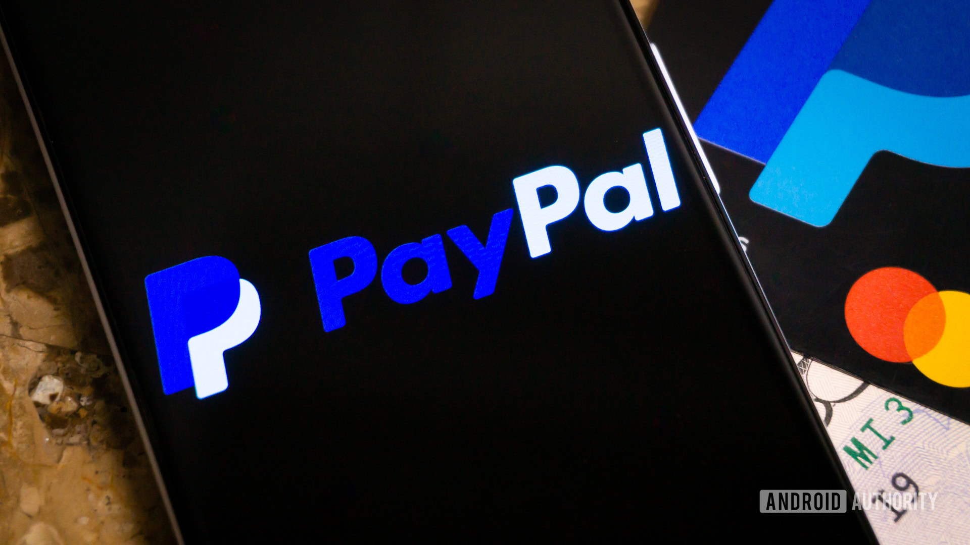 A stock photo of the PayPal logo shown on a smartphone lying on top of a credit card and some cash.