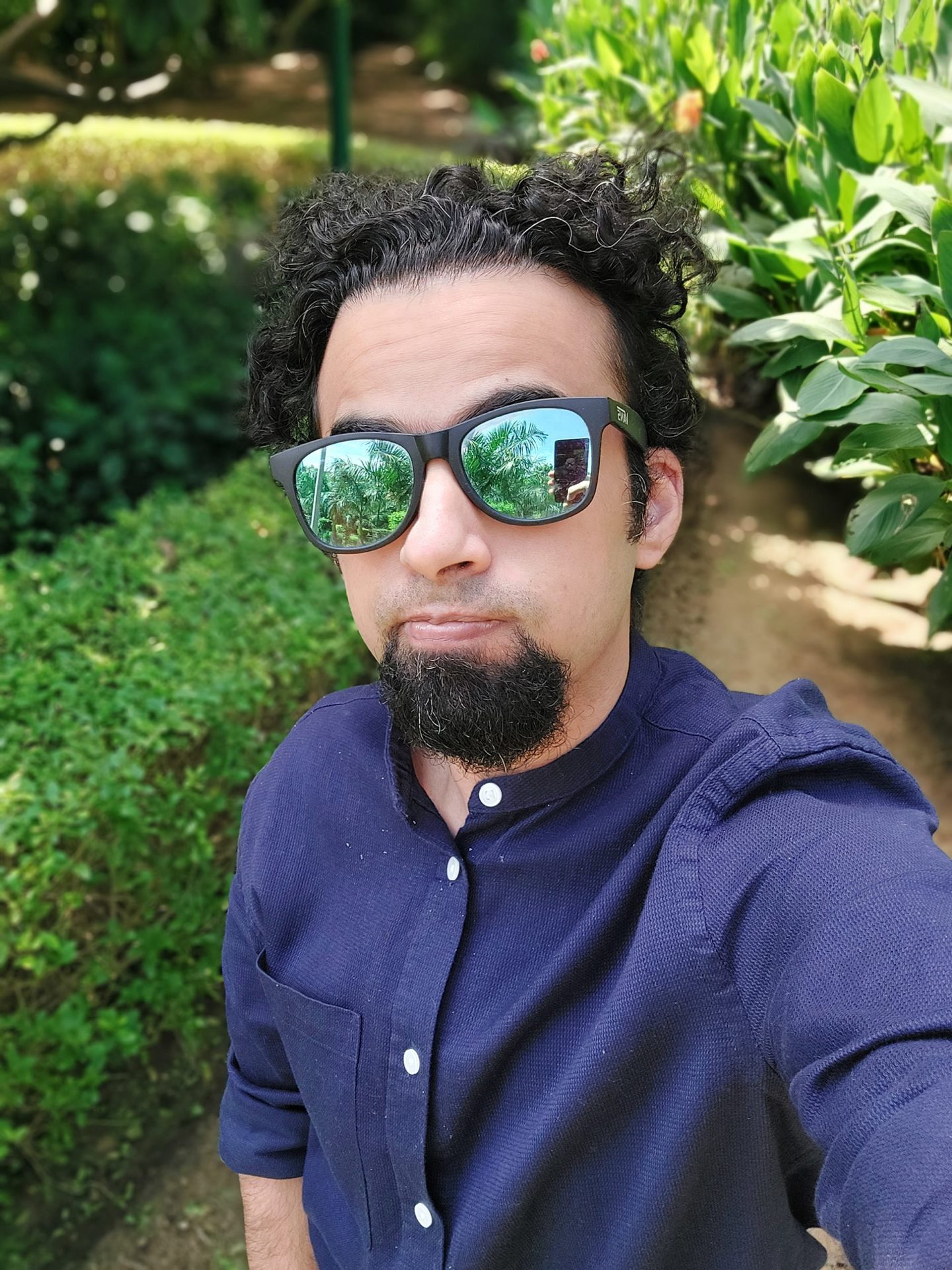 Samsung Galaxy A52s 5G selfie camera portrait mode shot of man with dark hair and beard wearing a blue long-sleeve shirt and mirrored shades, taken outdoors next to a green hedge.