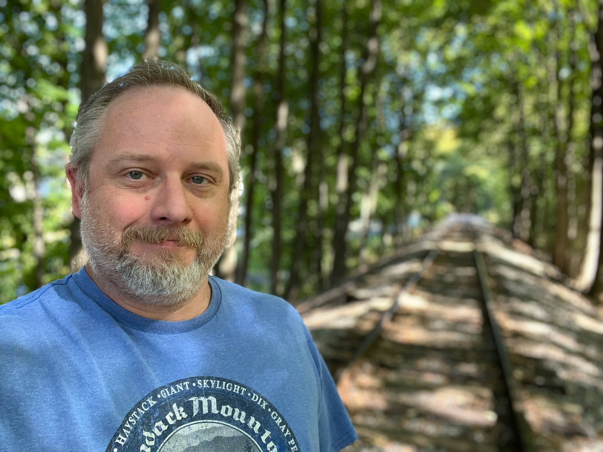 Apple iPhone 13 photo sample portrait on train tracks of a man with light colored hair and beard wearing a bright blue t-shirt
